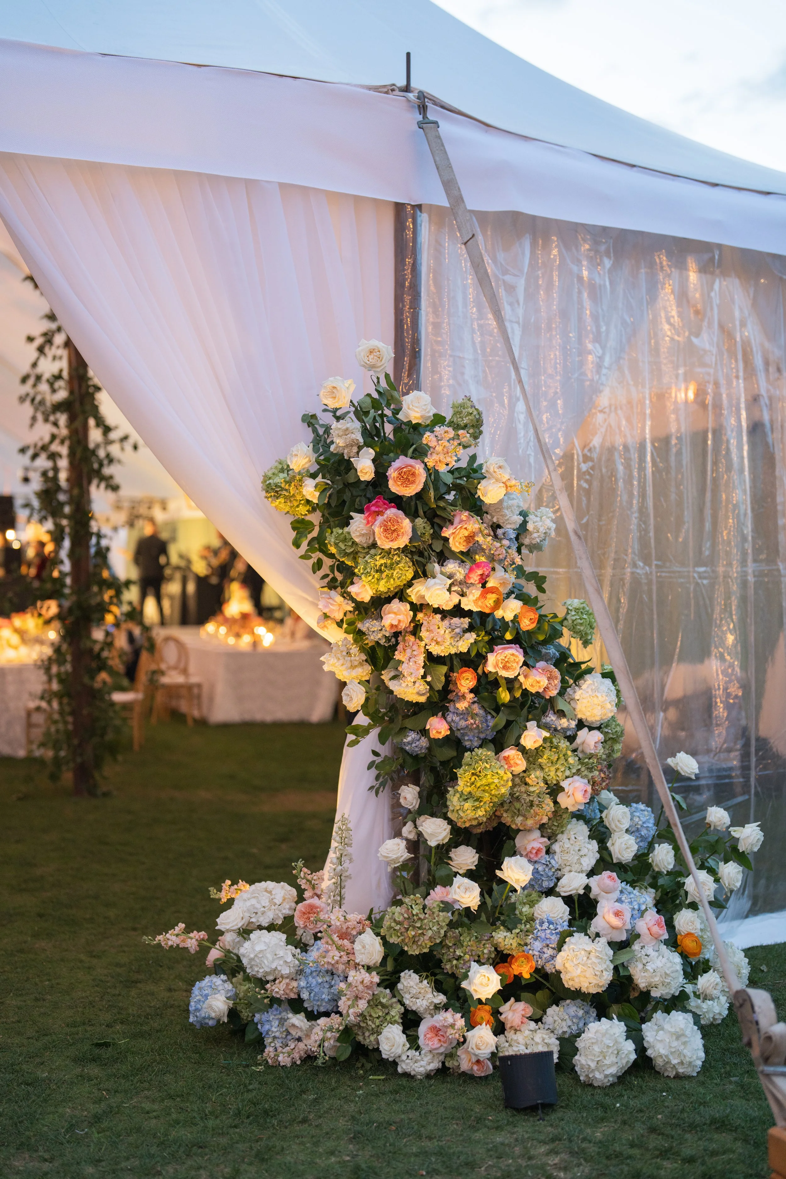Decorative floral arrangement with white, pink, and orange roses, and hydrangeas outside a white tent at a celebration.