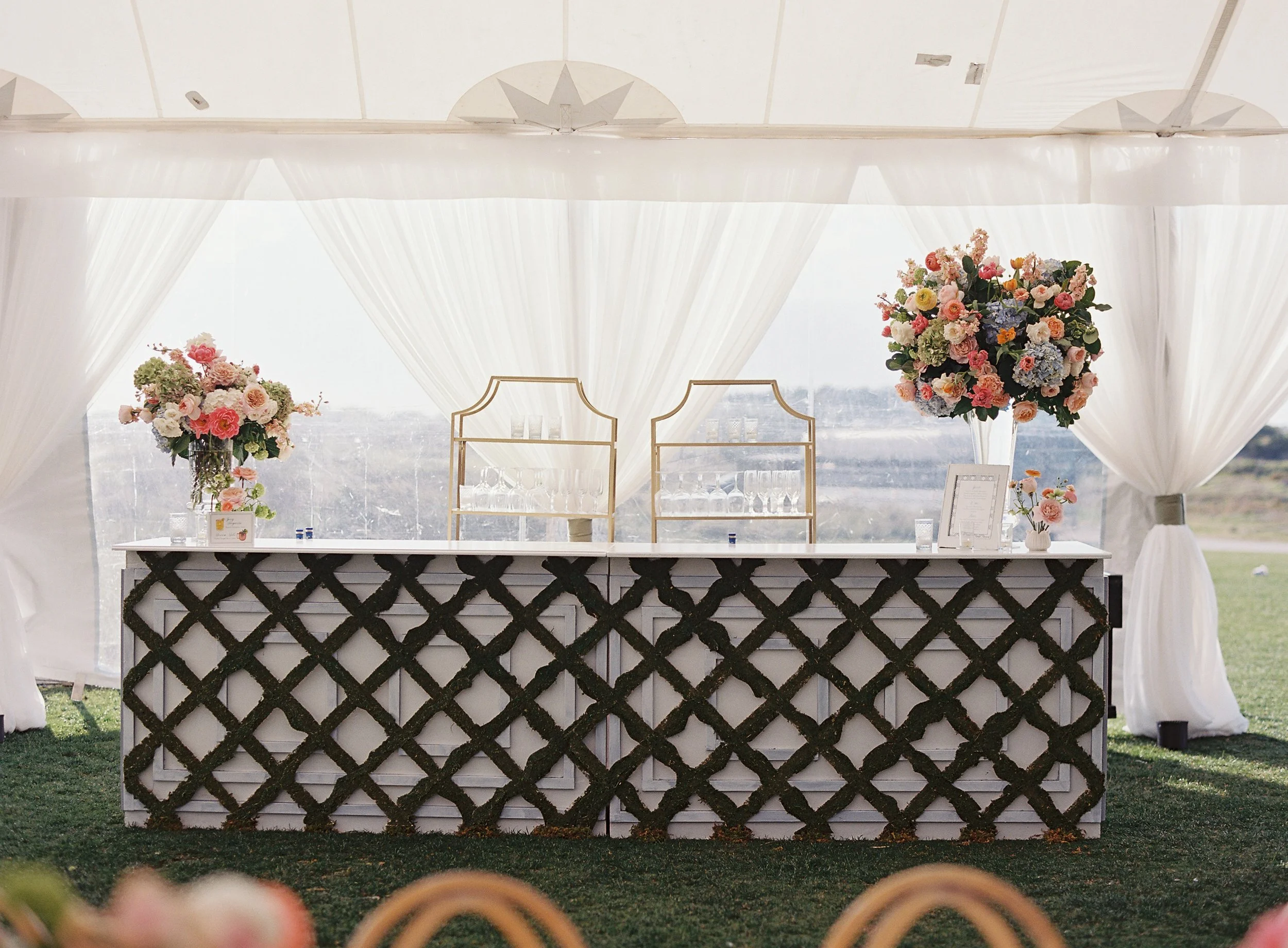 Elegant wedding bar setup with floral arrangements, champagne glasses, and a framed menu inside a white tent with draped curtains.