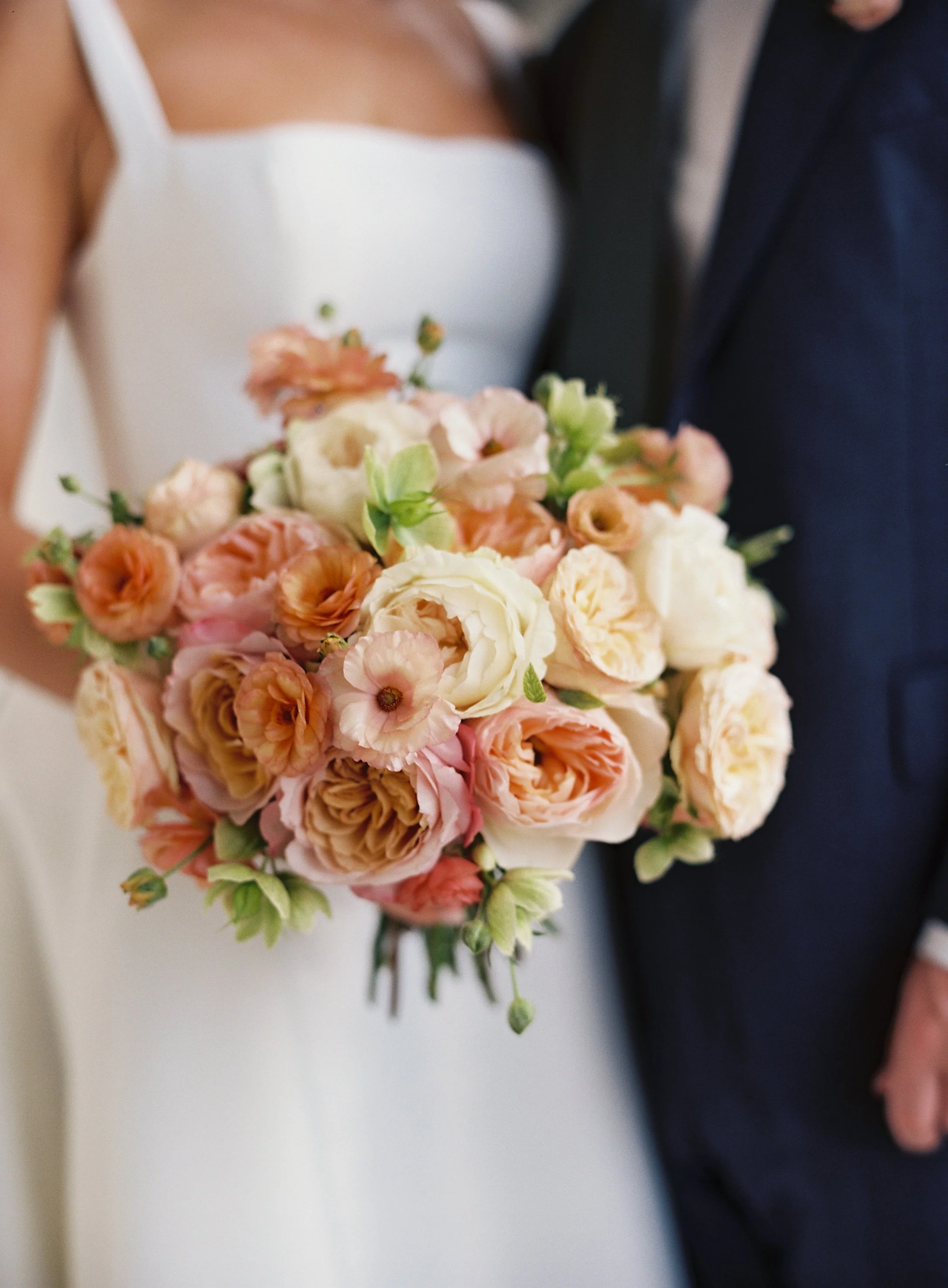 Close-up of a bride holding a bouquet of peach, white, and blush pink flowers, standing next to a groom in a dark suit on their wedding day at the Ocean Course on Kiawah Island, SC