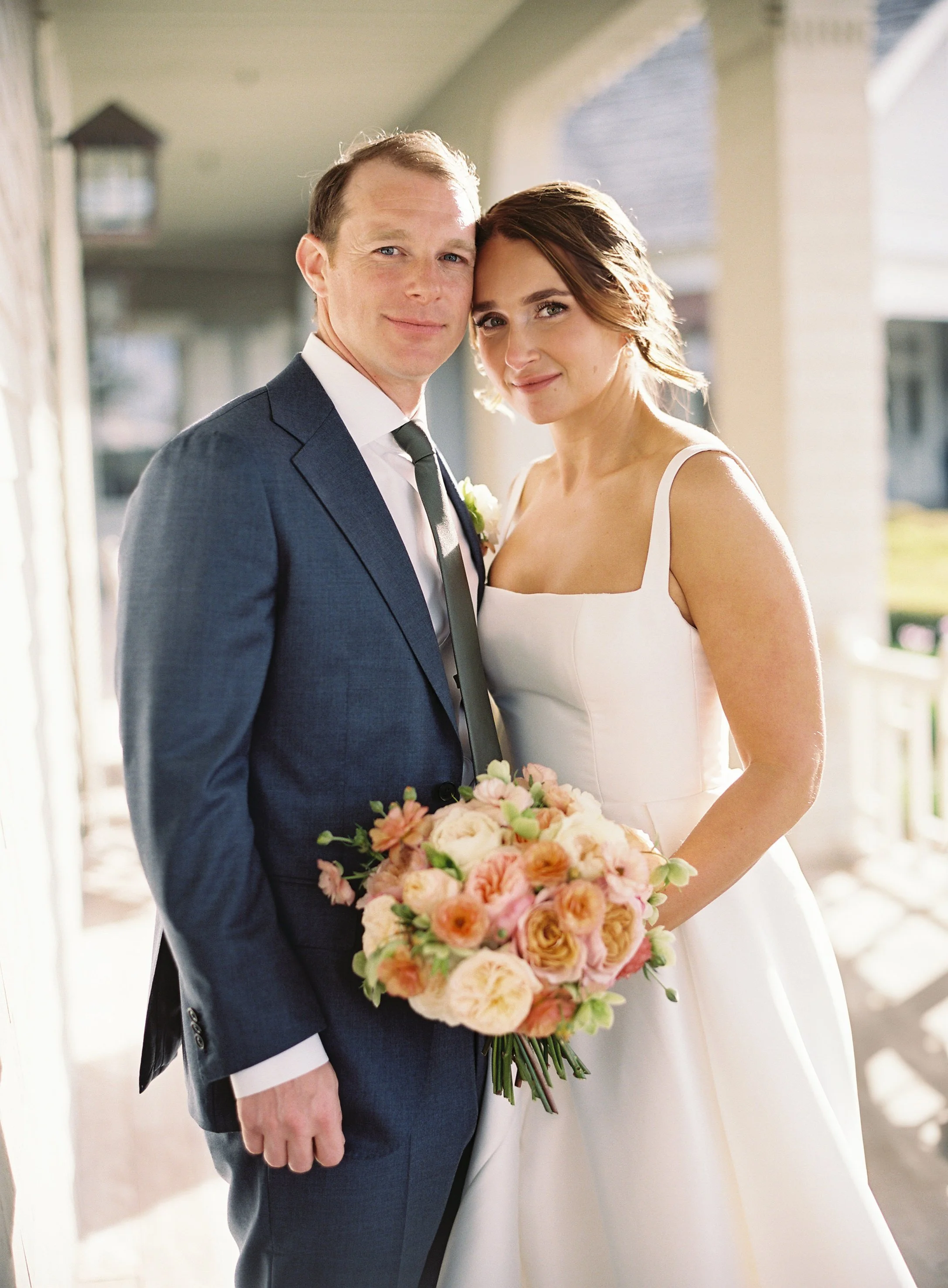 A bride and groom standing close together, smiling, with the bride holding a pink and peach bouquet, during a wedding at the Ocean Course on Kiawah Island, SC