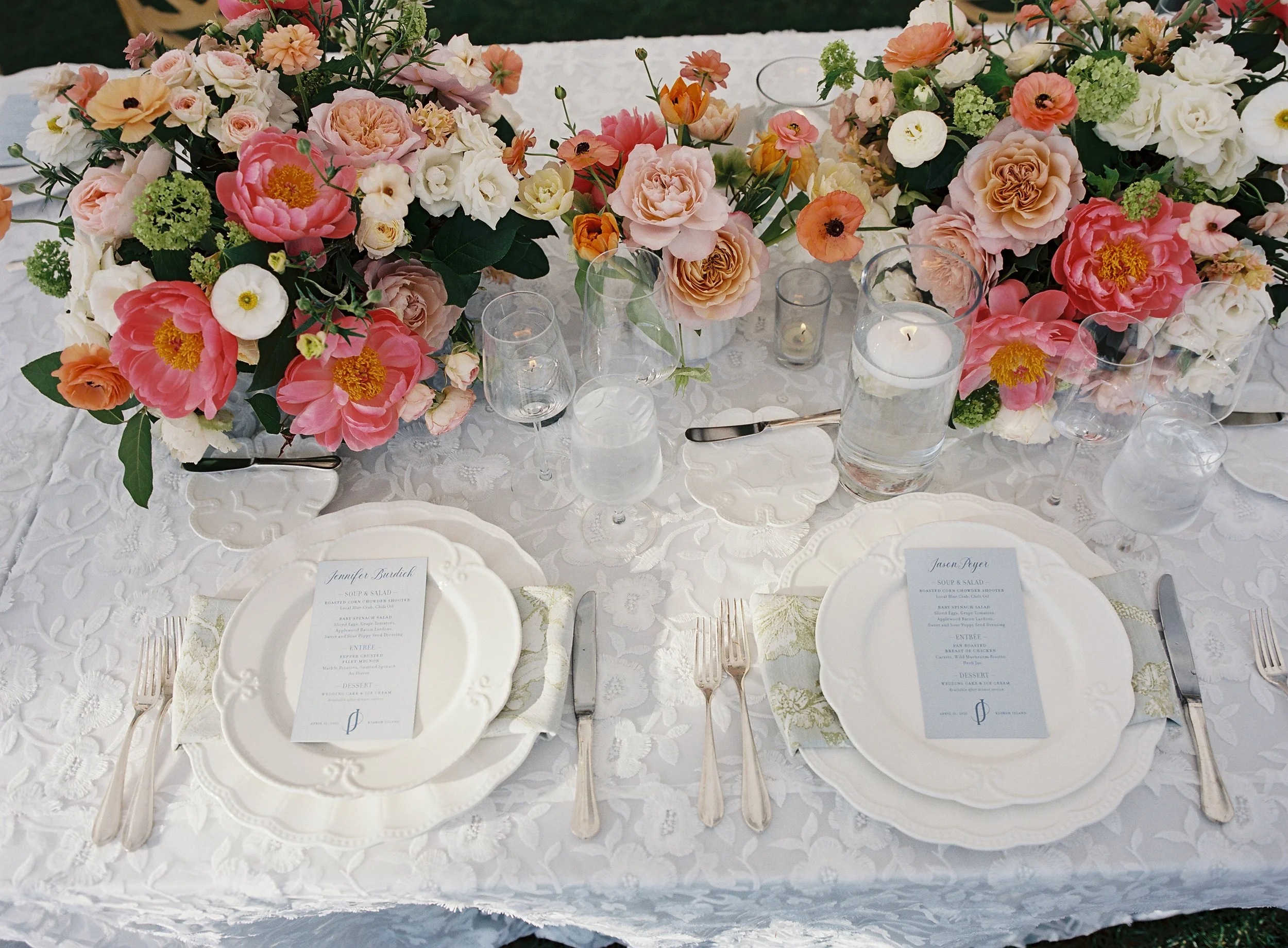 Elegant table setting with white lace tablecloth, floral centerpieces of pink, white, and peach roses with greenery, candles, and glassware. Each place has a white plate, floral-patterned napkin, menu, and silverware.