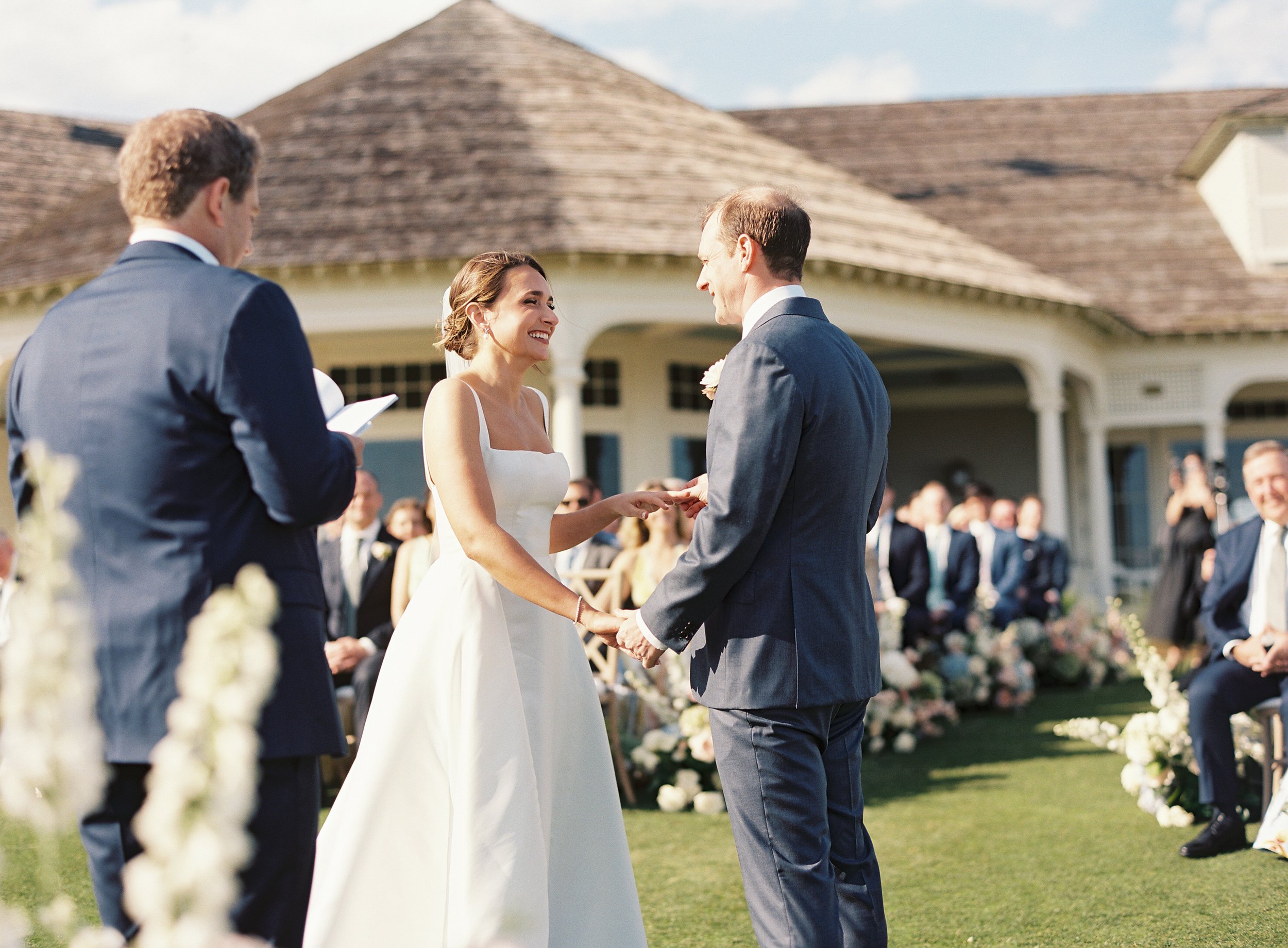 A bride and groom exchanging vows at an outdoor wedding ceremony, with guests seated in the background.