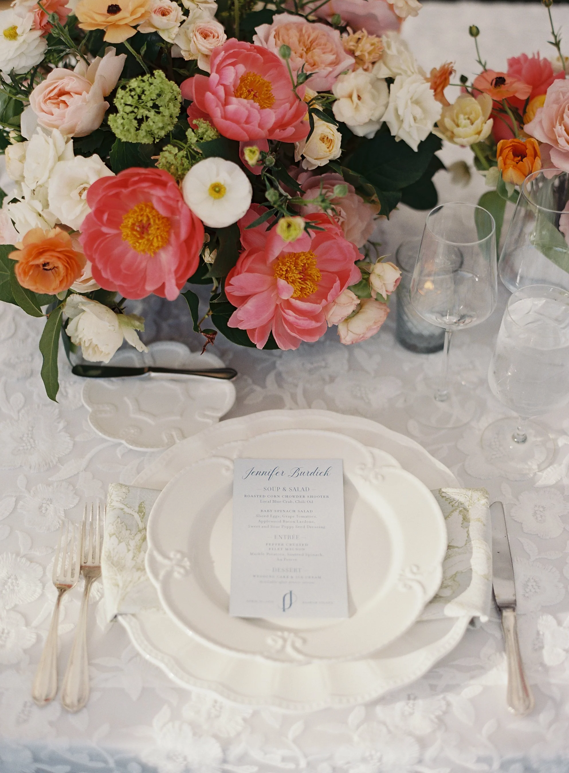 Elegant table setting with a floral centerpiece of pink, white, and peach flowers, a white plate with a menu card, silverware, a wine glass, a water glass, and decorative linens.
