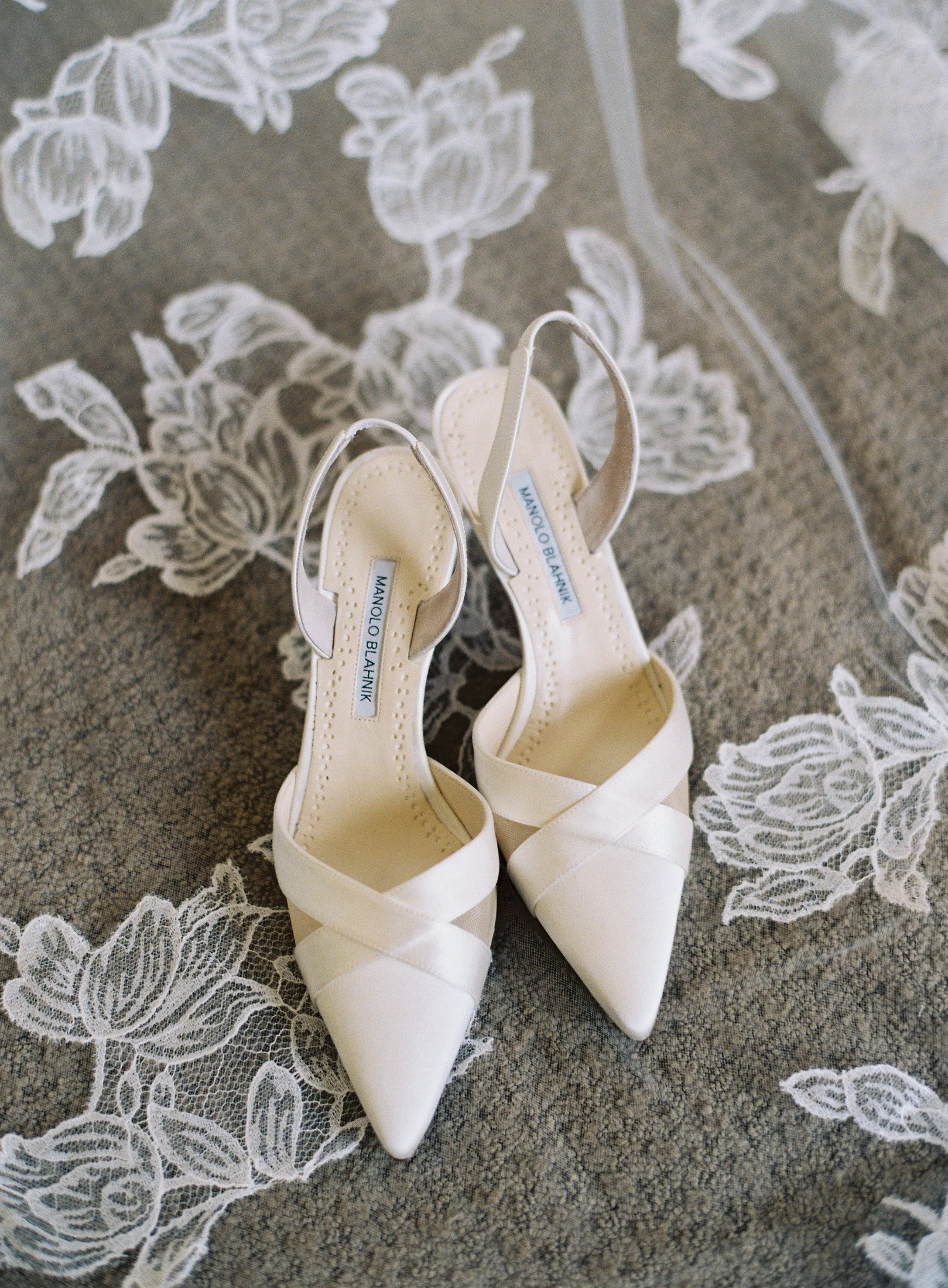 A pair of white Manolo Blahnik high-heeled shoes with pointed toes and crossed straps, placed on a gray fabric with white lace floral embroidery.