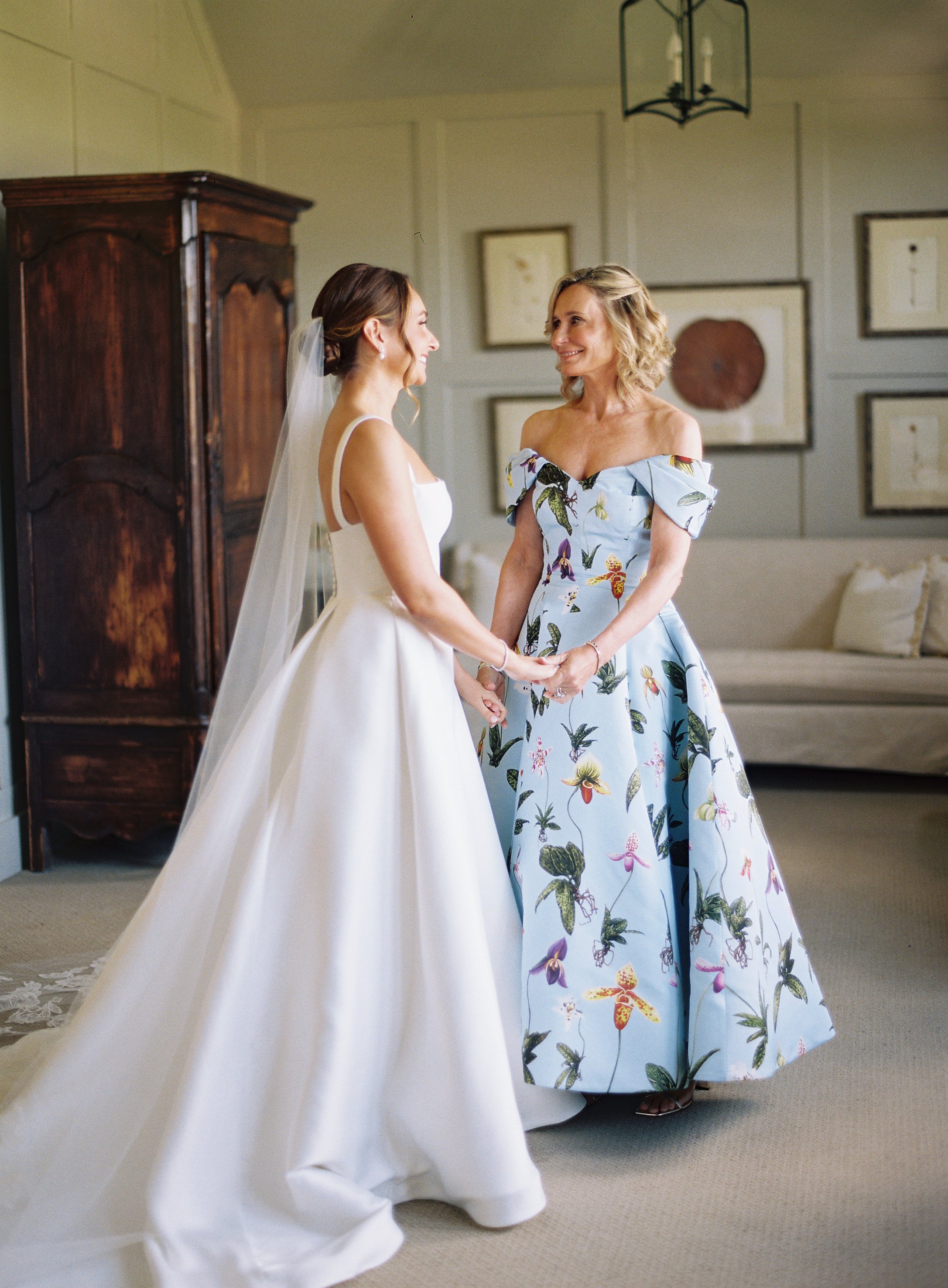 A bride in a white wedding dress holding hands with a woman in a floral off-the-shoulder dress, both smiling and looking at each other inside a decorated room.