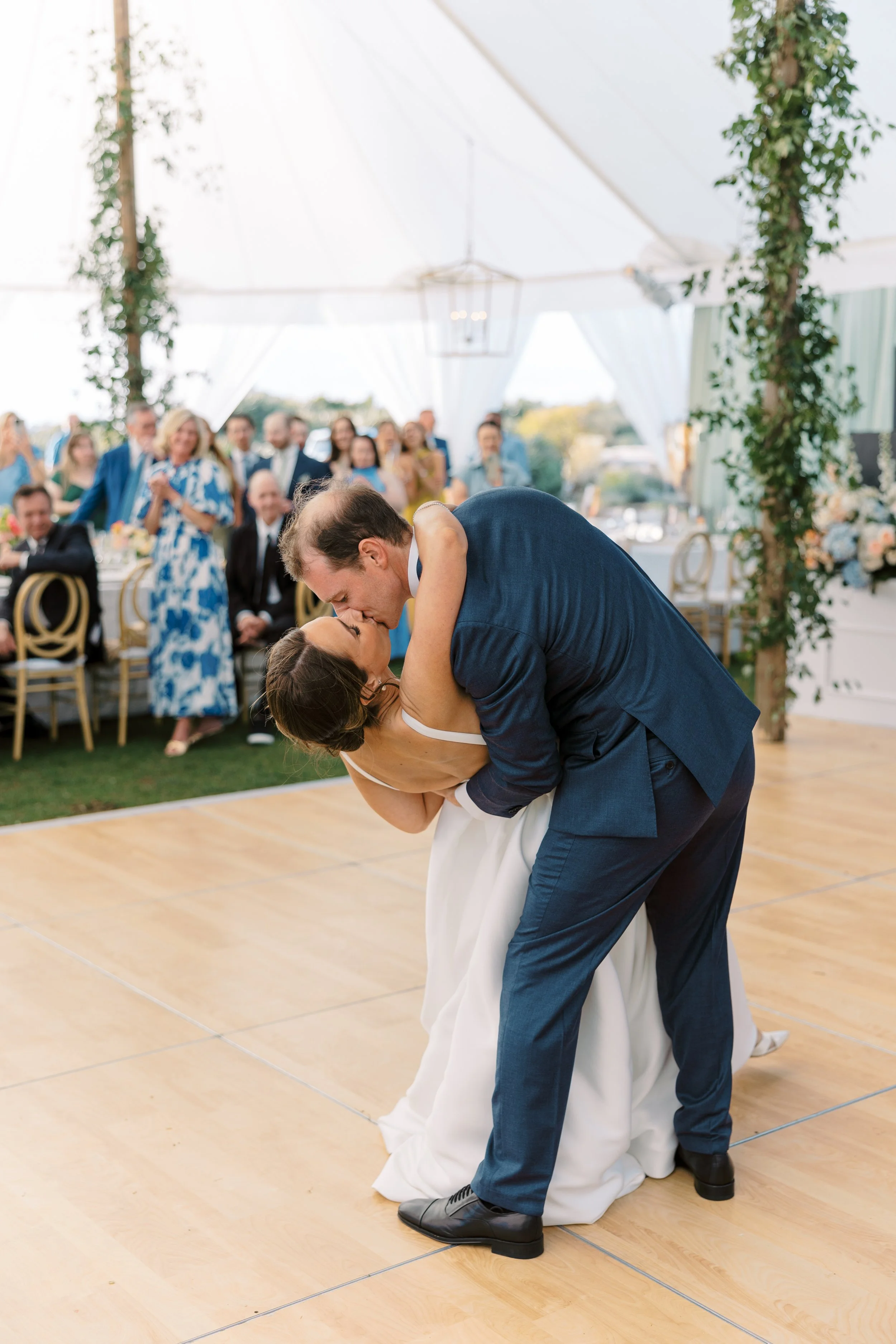 A bride and groom sharing a kiss during their wedding dance under a large white tent with wedding guests watching in the background.