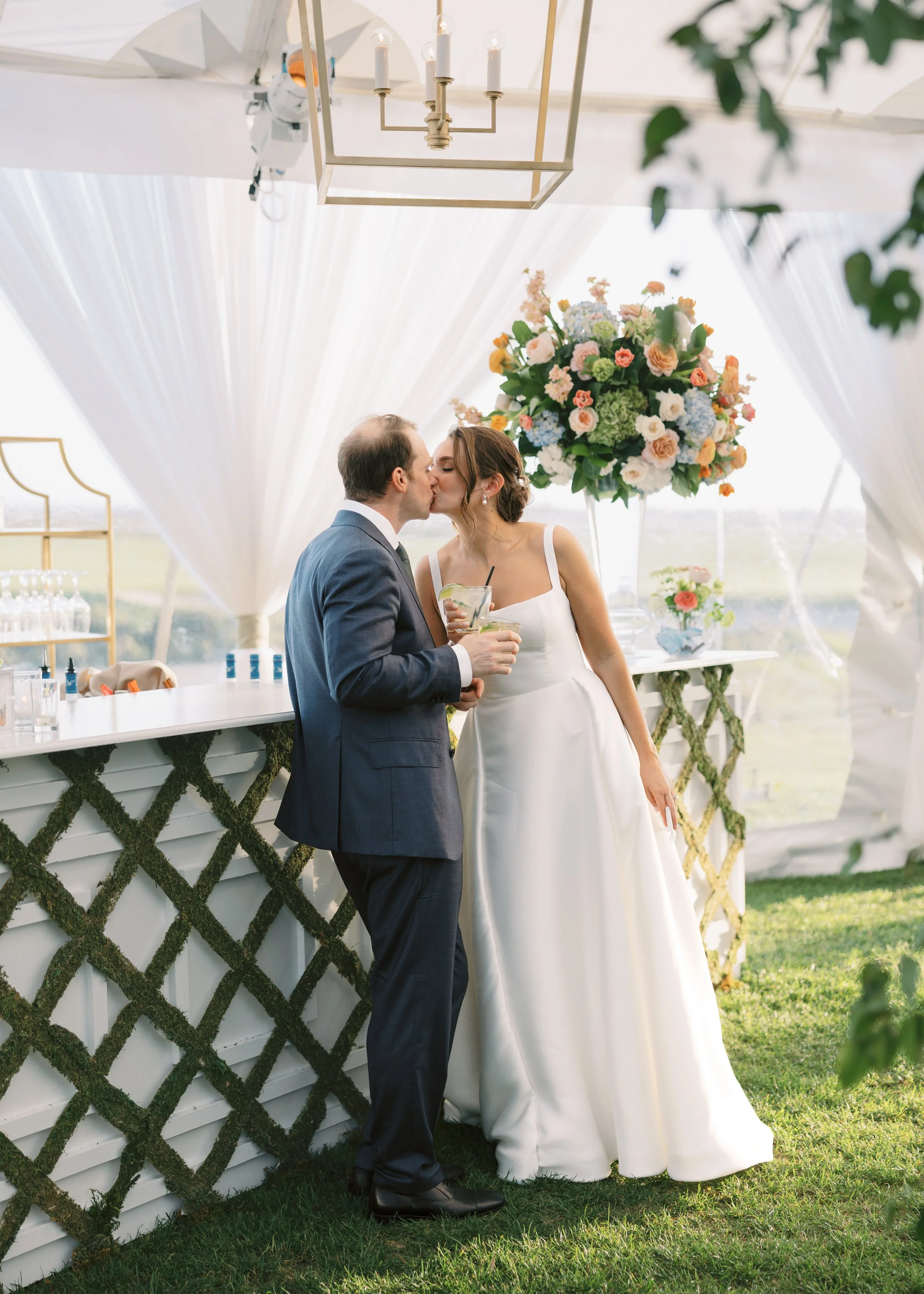 A bride and groom sharing a kiss at their wedding reception, with drinks in hand, under a decorated white canopy with floral arrangements and a chandelier overhead.