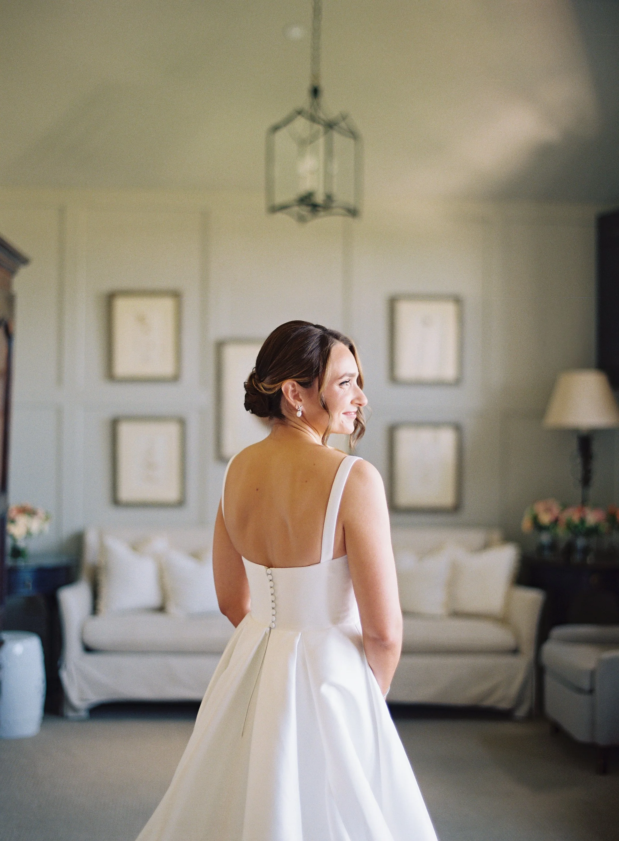 A bride with light skin, wearing a white wedding dress with buttons down the back, stands in a living room with a white sofa, framed artwork, and soft lighting, smiling as she looks to the side.