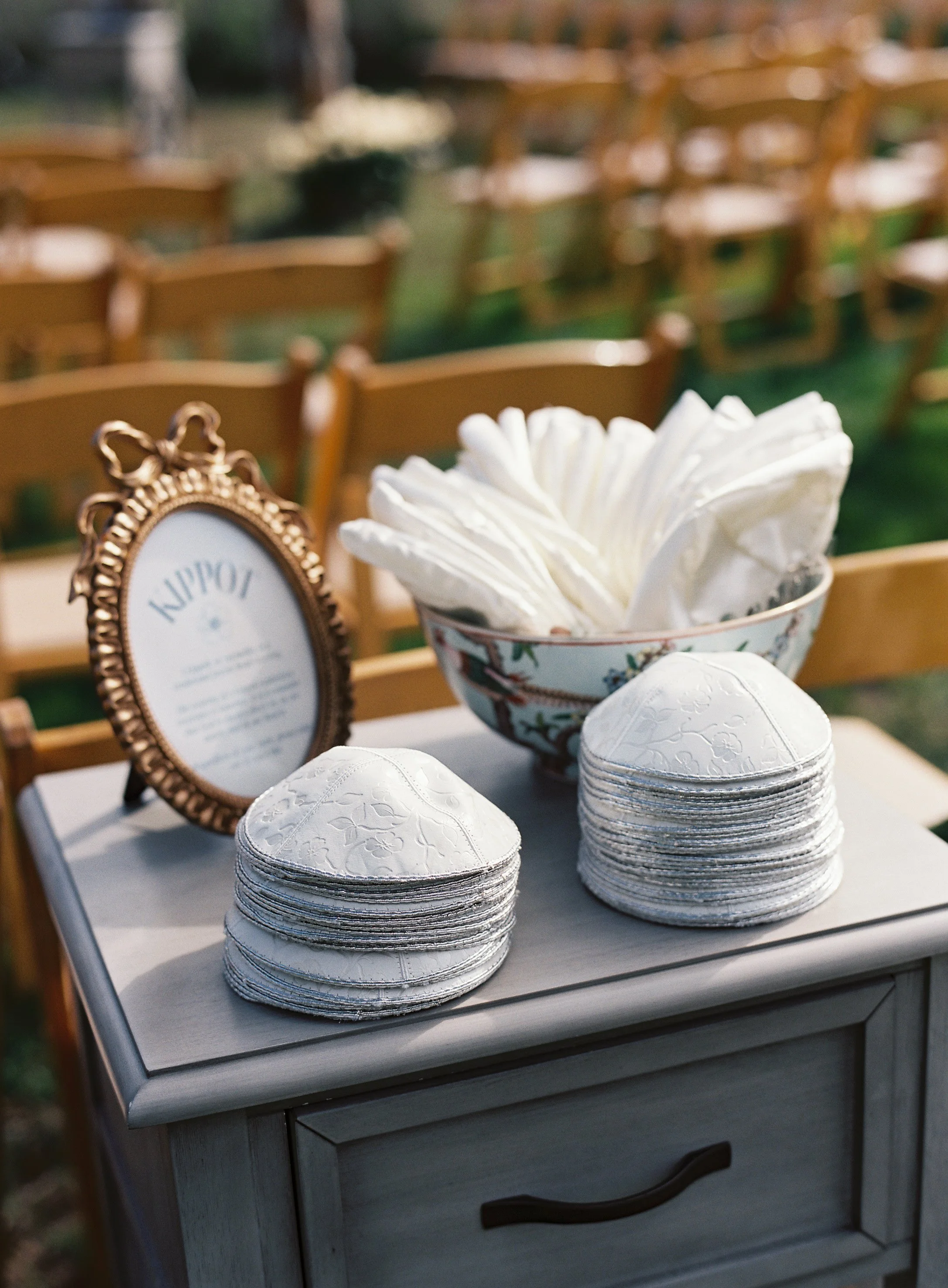 A table set up outdoors with paper napkins, a bowl of white napkins, and a decorative framed sign, surrounded by multiple wooden chairs.