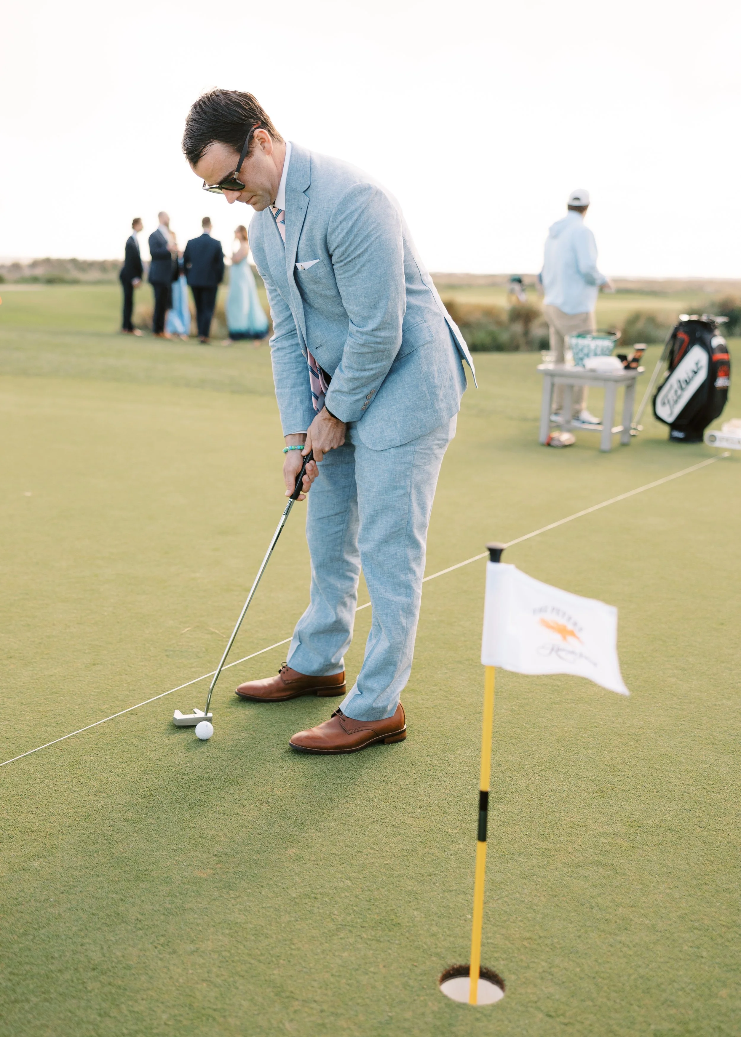 A man in a light gray suit and sunglasses prepares to putt on a golf course, with a group of people dressed in formal attire in the background.