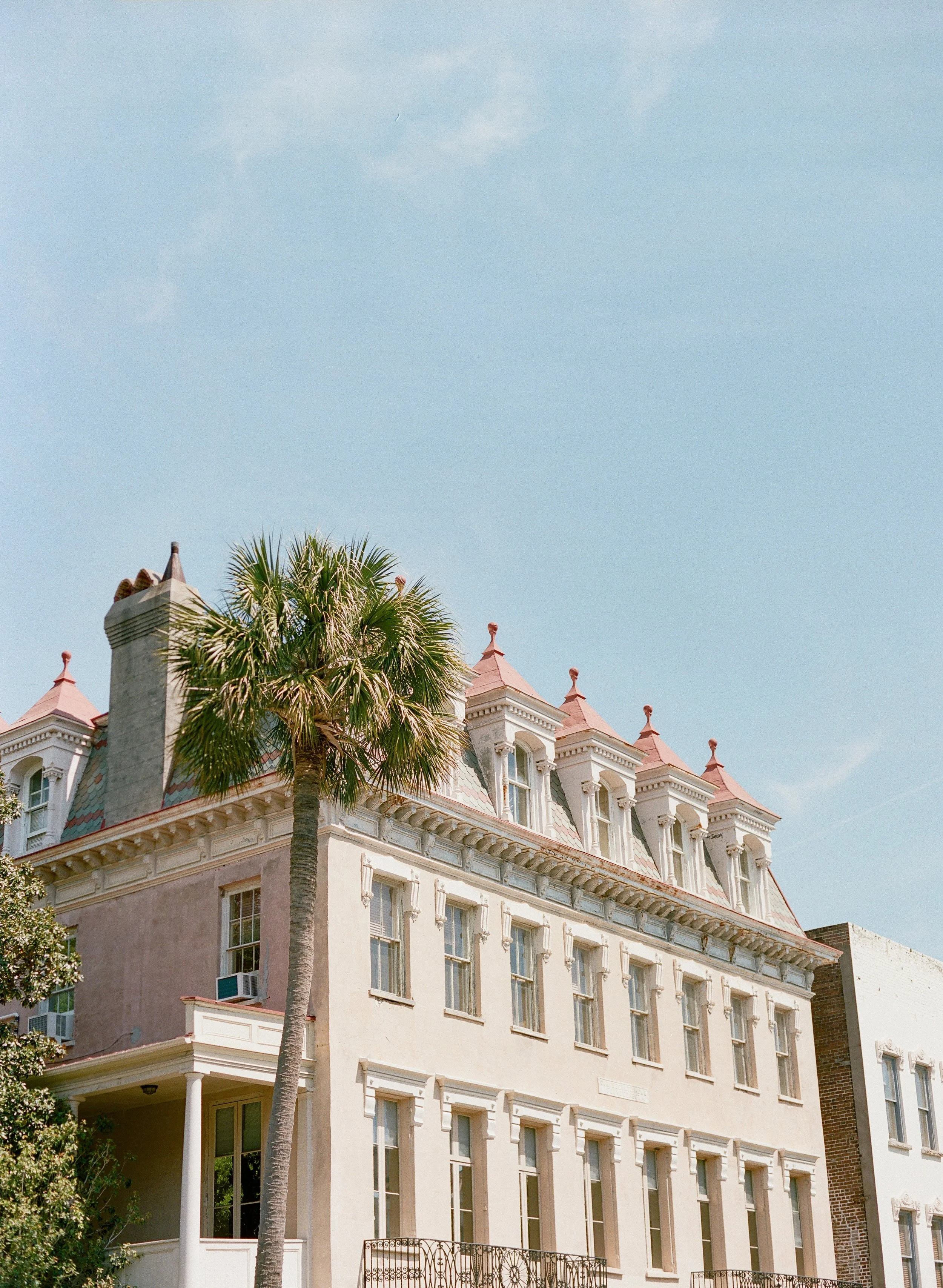 A multi-story building in Charleston, South Carolina, with a pale exterior, ornate architectural details, and a steeply pitched roof with dormer windows. A tall palm tree stands in front of the building against a clear blue sky.