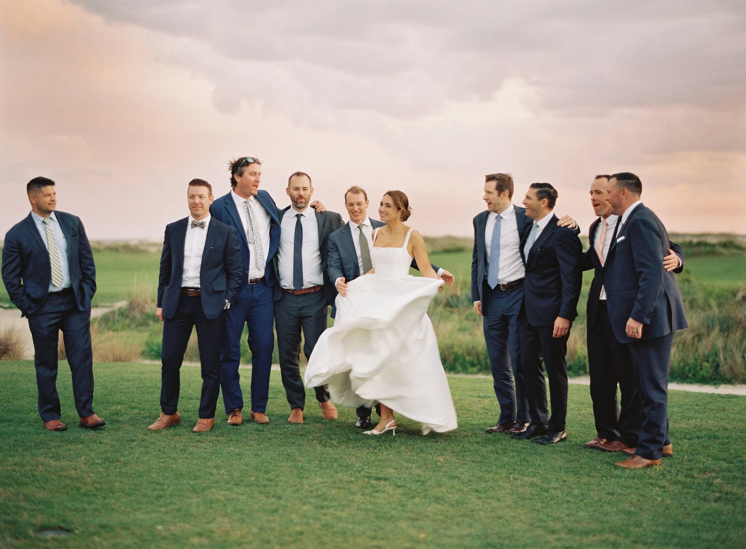 A bride in a white wedding dress poses with a group of men in suits outdoors on a grassy field at sunset.