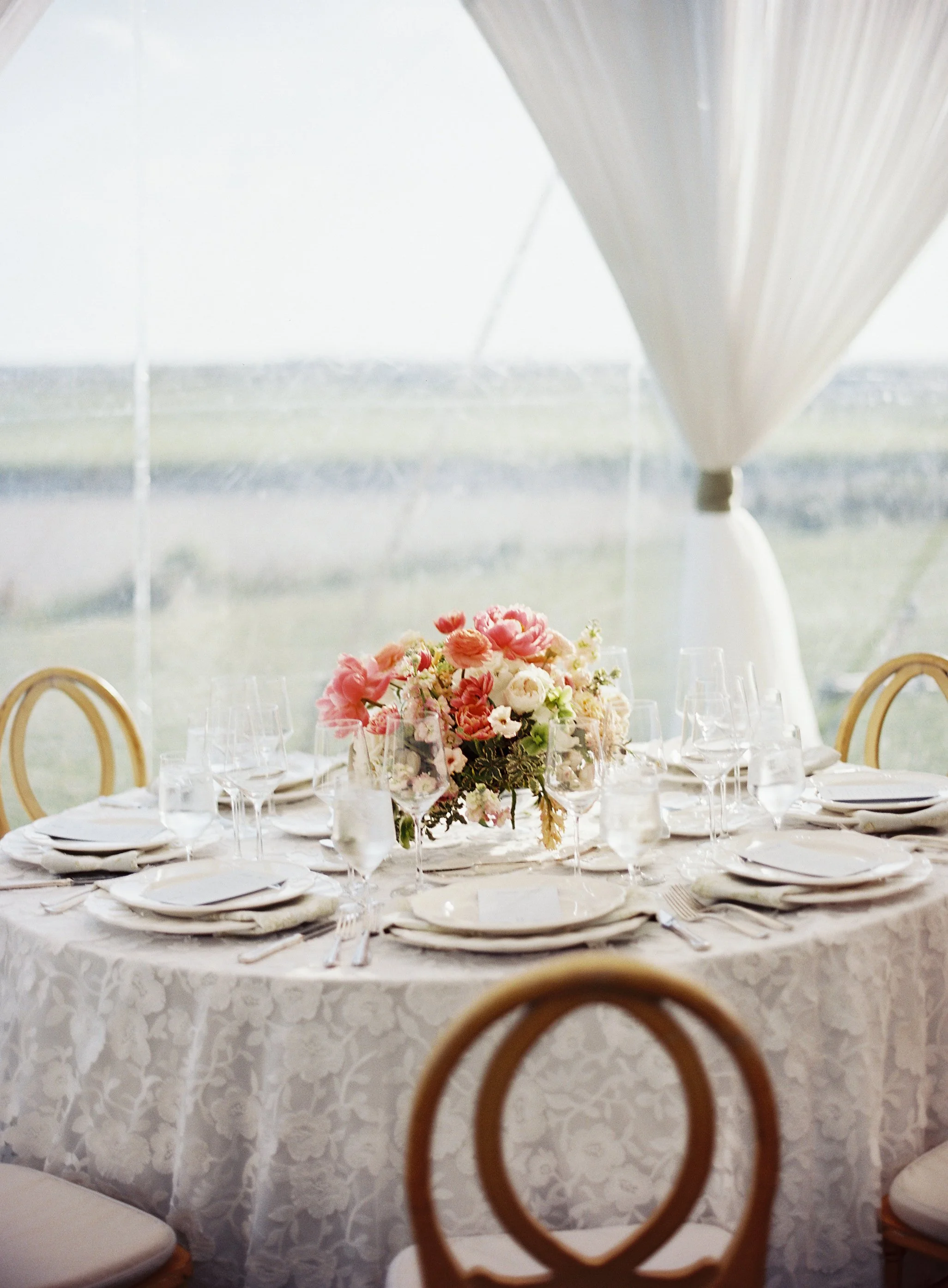 Round banquet table set for a formal event with a floral centerpiece, multiple wine glasses, and neatly arranged plates and silverware, under a white canopy with sunlight streaming in.