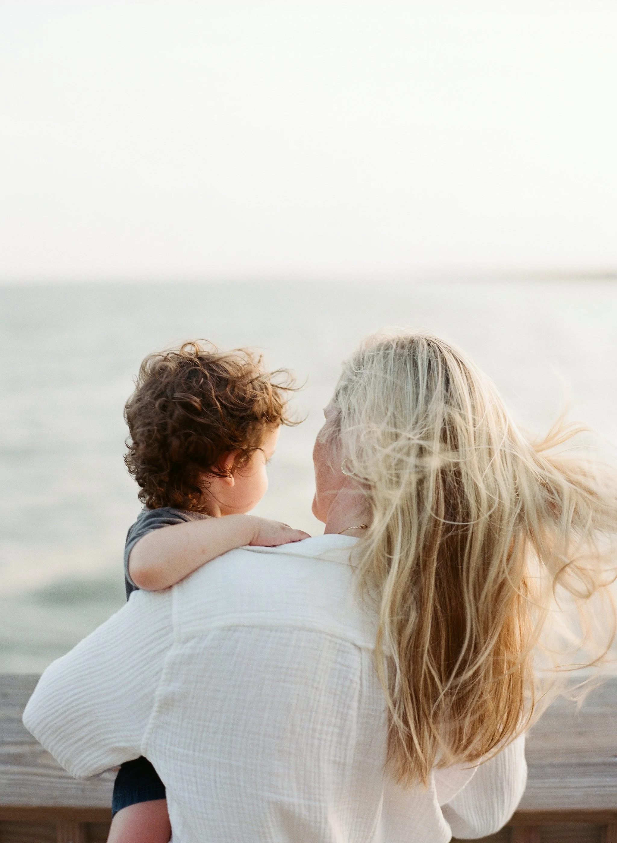 Portrait of Lauren Jonas from behind holding her young child by the water at the beach.