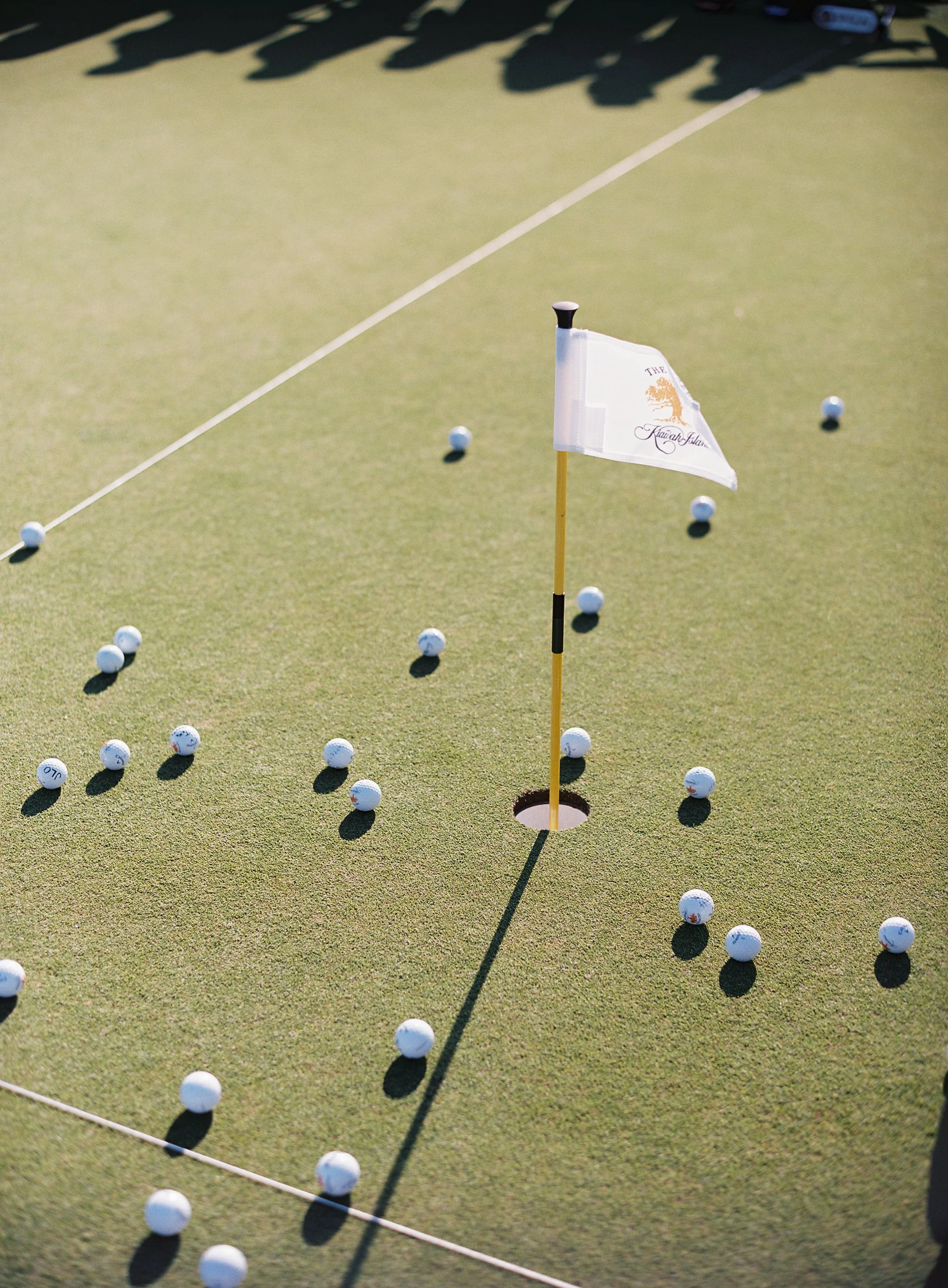 Golf green with many golf balls scattered around, a flagstick in the hole, and shadows of trees or players in the background.