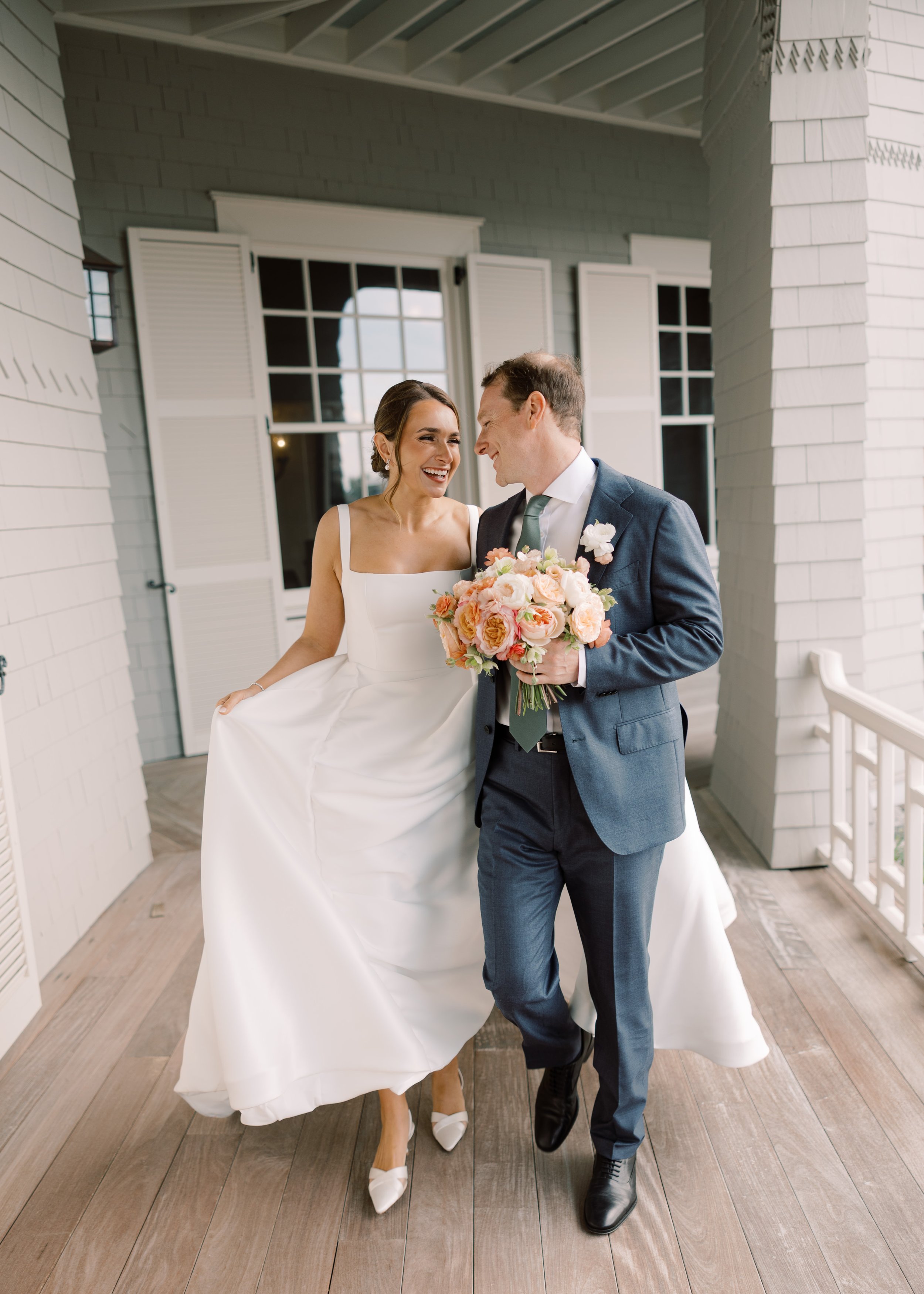A bride and groom smiling at each other on a porch, the bride holding up her wedding dress and the groom holding a bouquet of pink and peach flowers.