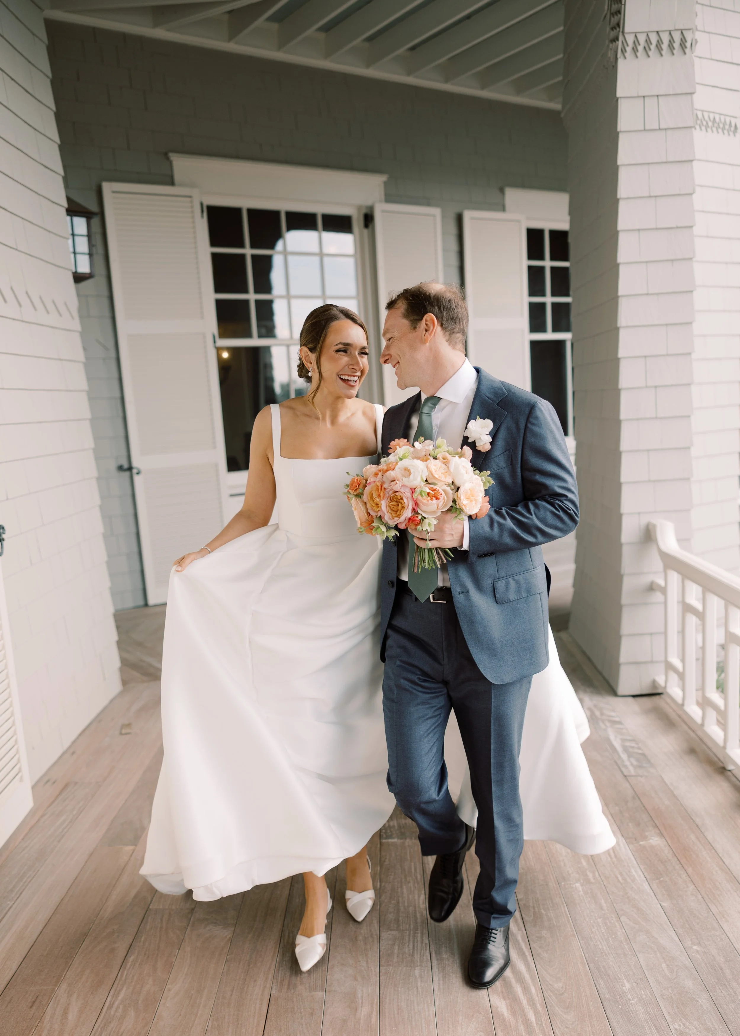 A bride and groom smiling at each other on a porch, the bride holding up her wedding dress and the groom holding a bouquet of pink and peach flowers.