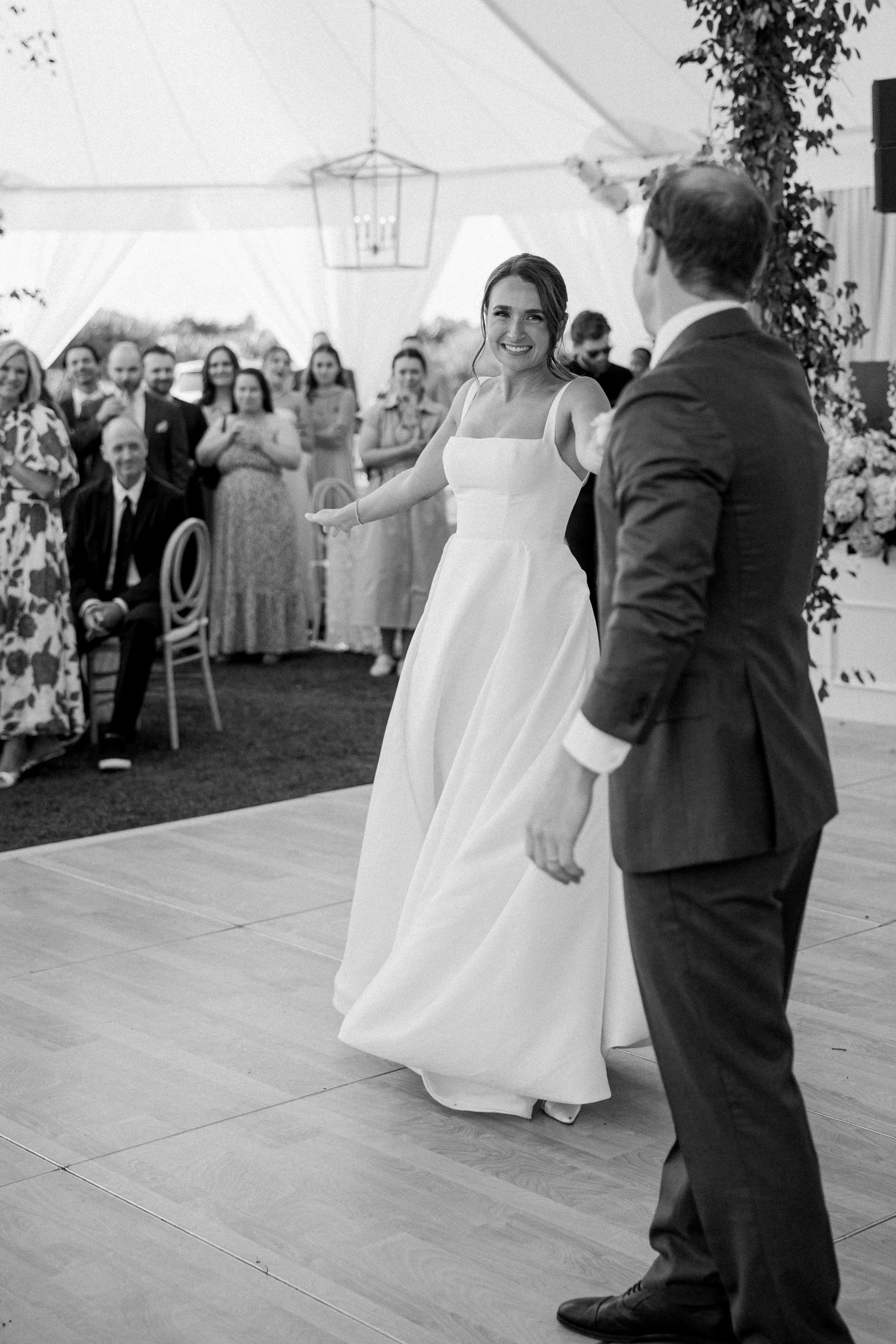 A bride in a white wedding dress smiling and dancing with a groom in a suit during a wedding reception under a tent, with guests watching in the background.