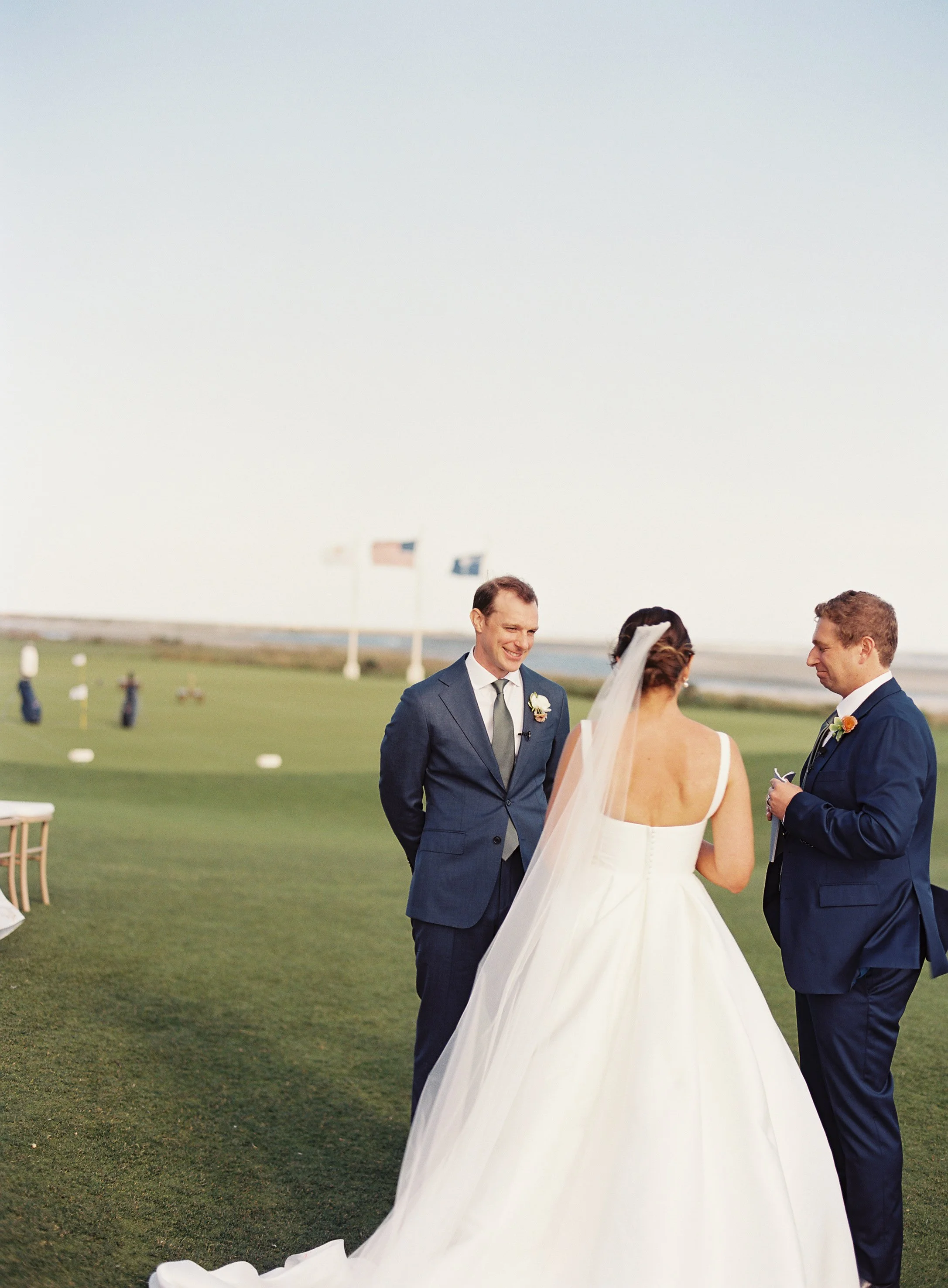 A wedding ceremony taking place outdoors on a grassy field, with a bride in a white gown and veil facing two men in suits, one smiling, as they exchange vows or rings. In the background, flags and a body of water are visible.