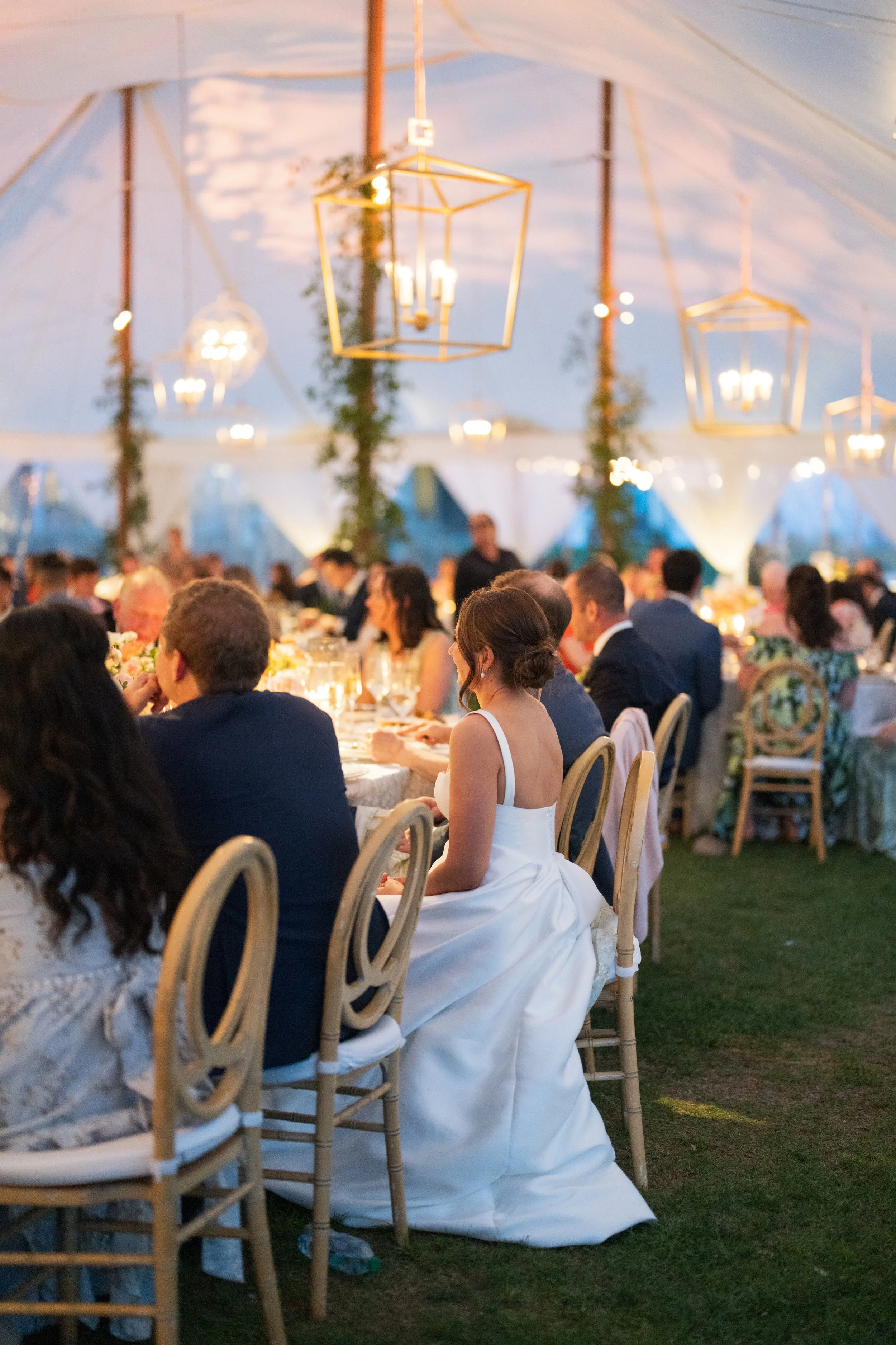 Elegant wedding reception under a tent with hanging geometric chandeliers, guests seated at tables, and a bride in a white gown sitting among wedding guests.