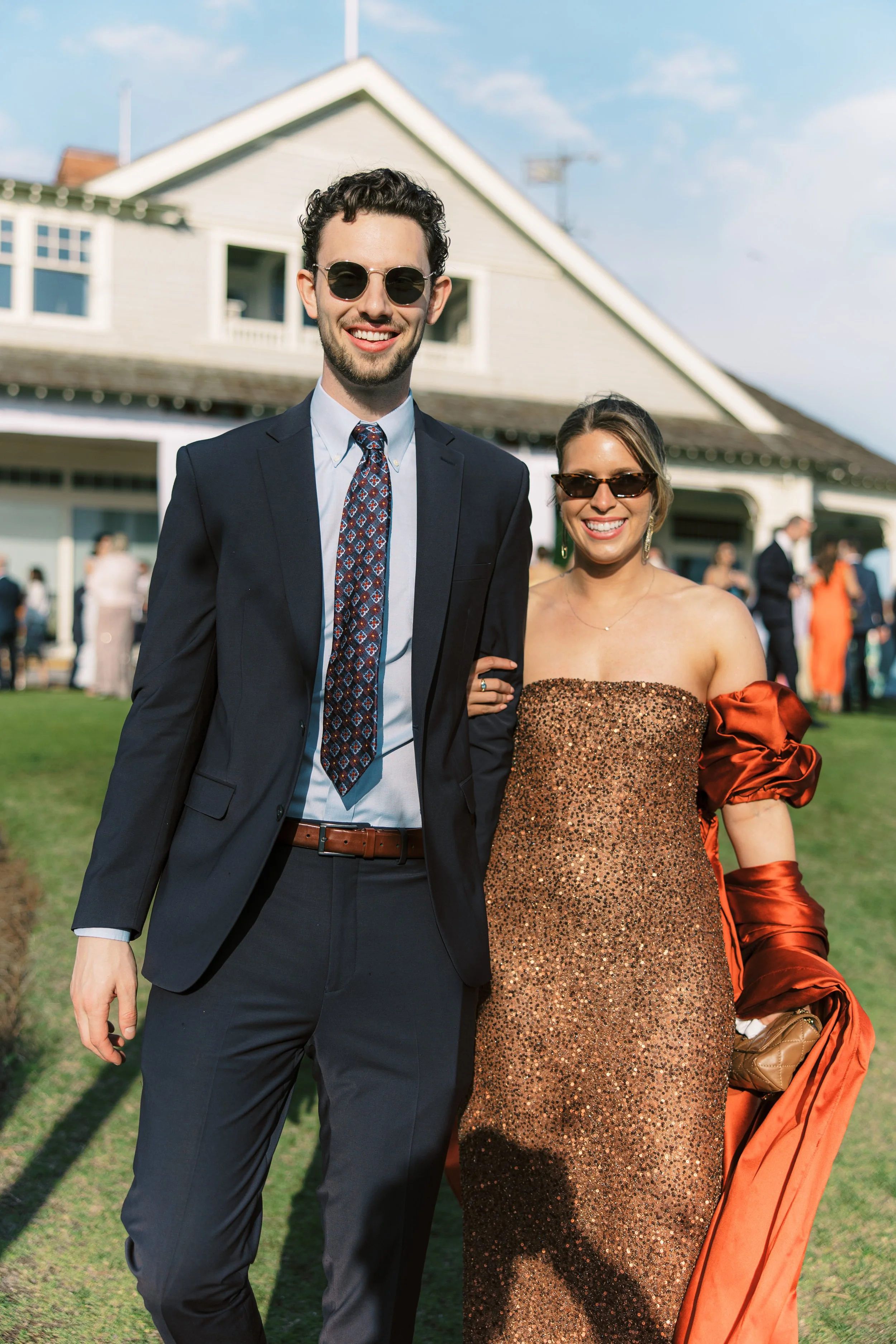 A man and a woman dressed in formal attire standing outdoors at a social event, with a house in the background and other guests mingling.