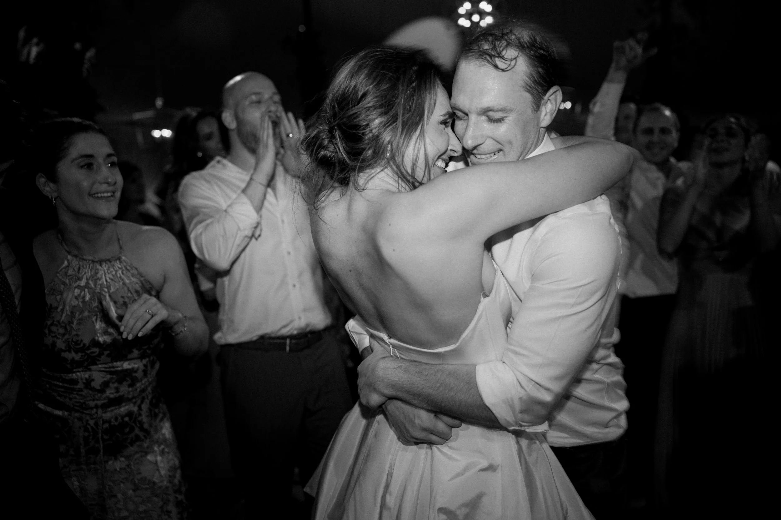 A black and white photo of a joyful bride and groom embracing each other during their wedding dance, surrounded by happy guests.