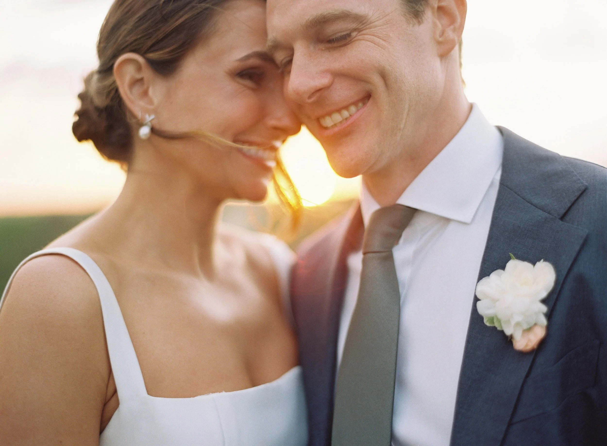 A happy bride and groom close together, smiling at each other outdoors during sunset.