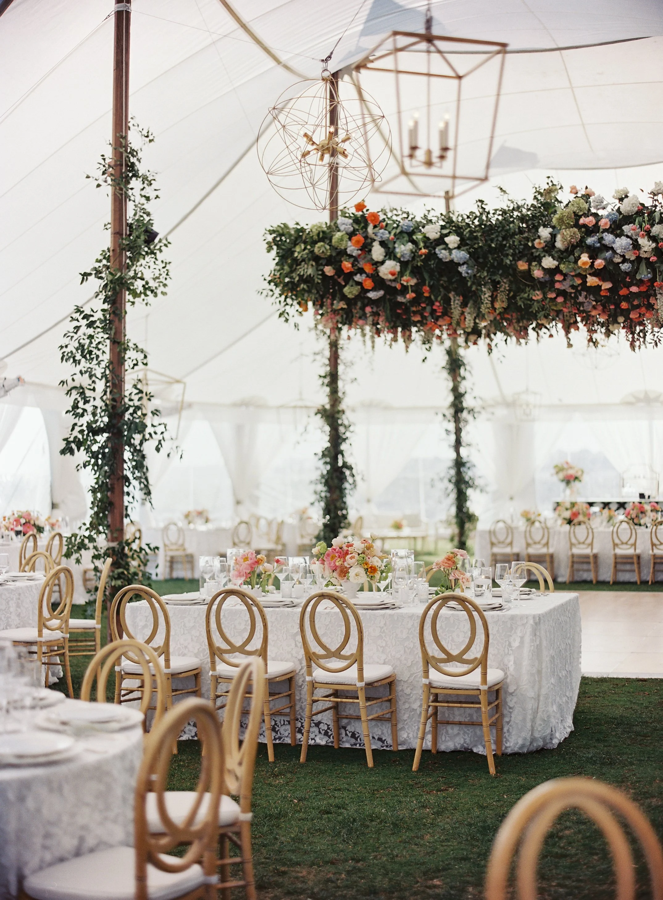Elegant wedding or event reception setup inside a large tent with decorated tables, floral arrangements, and hanging lights.