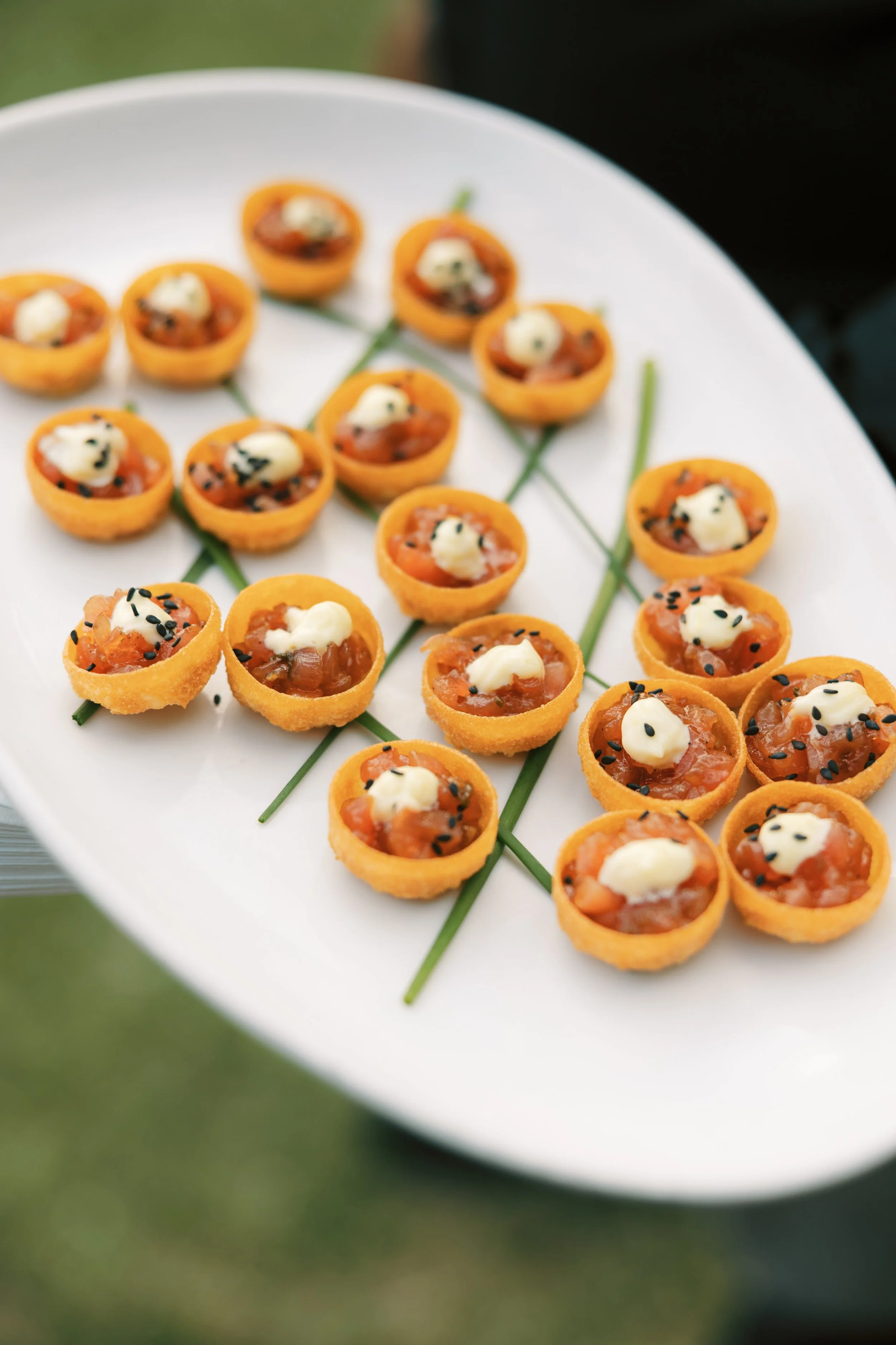 Miniature savory tartlets topped with cream, black sesame seeds, and diced vegetables, arranged on a white oval plate with green chive garnish during a cocktail hour in Charleston, SC 