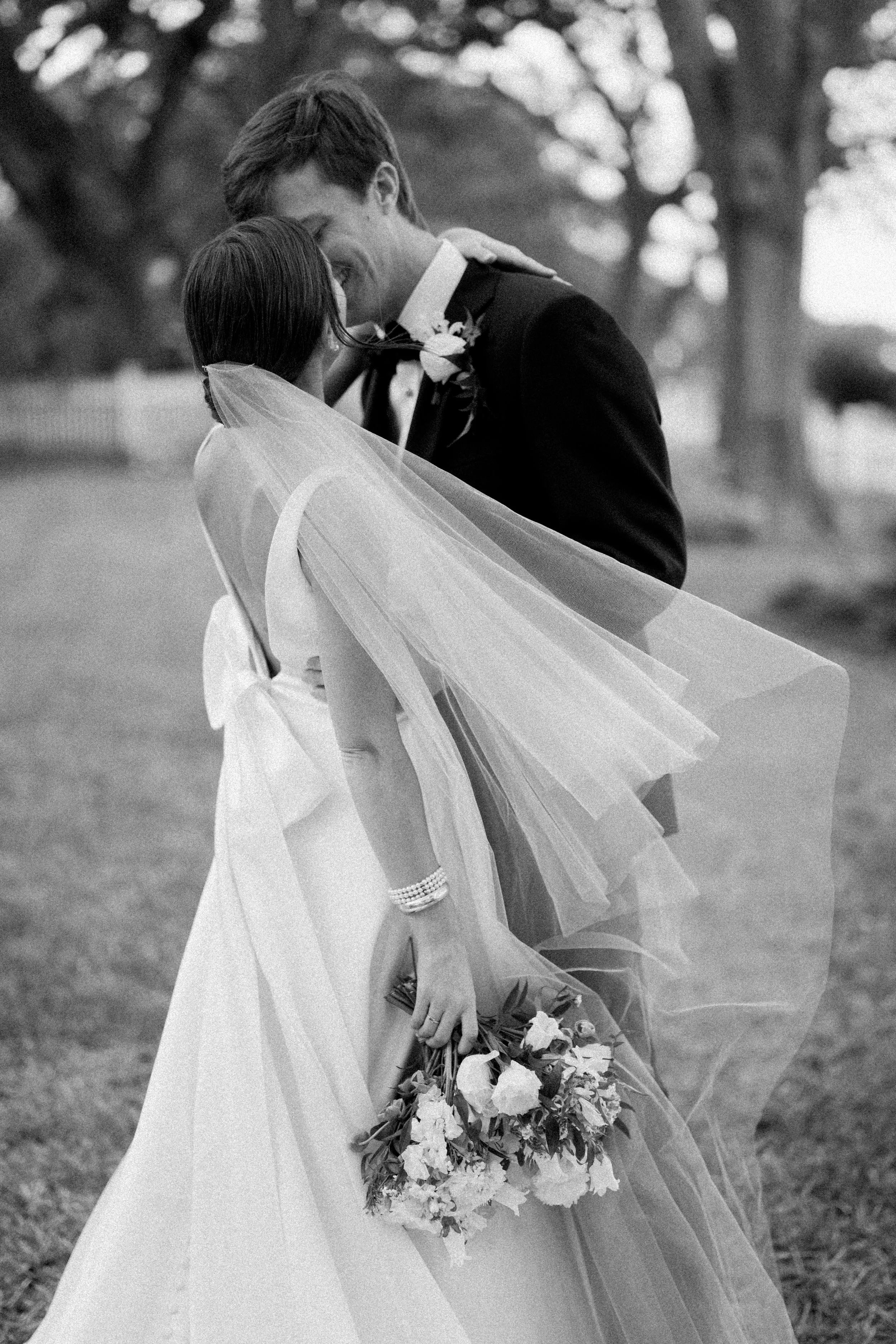 A black and white photo of a bride and groom in wedding attire sharing a close, intimate moment outdoors, with the bride holding a bouquet of flowers.