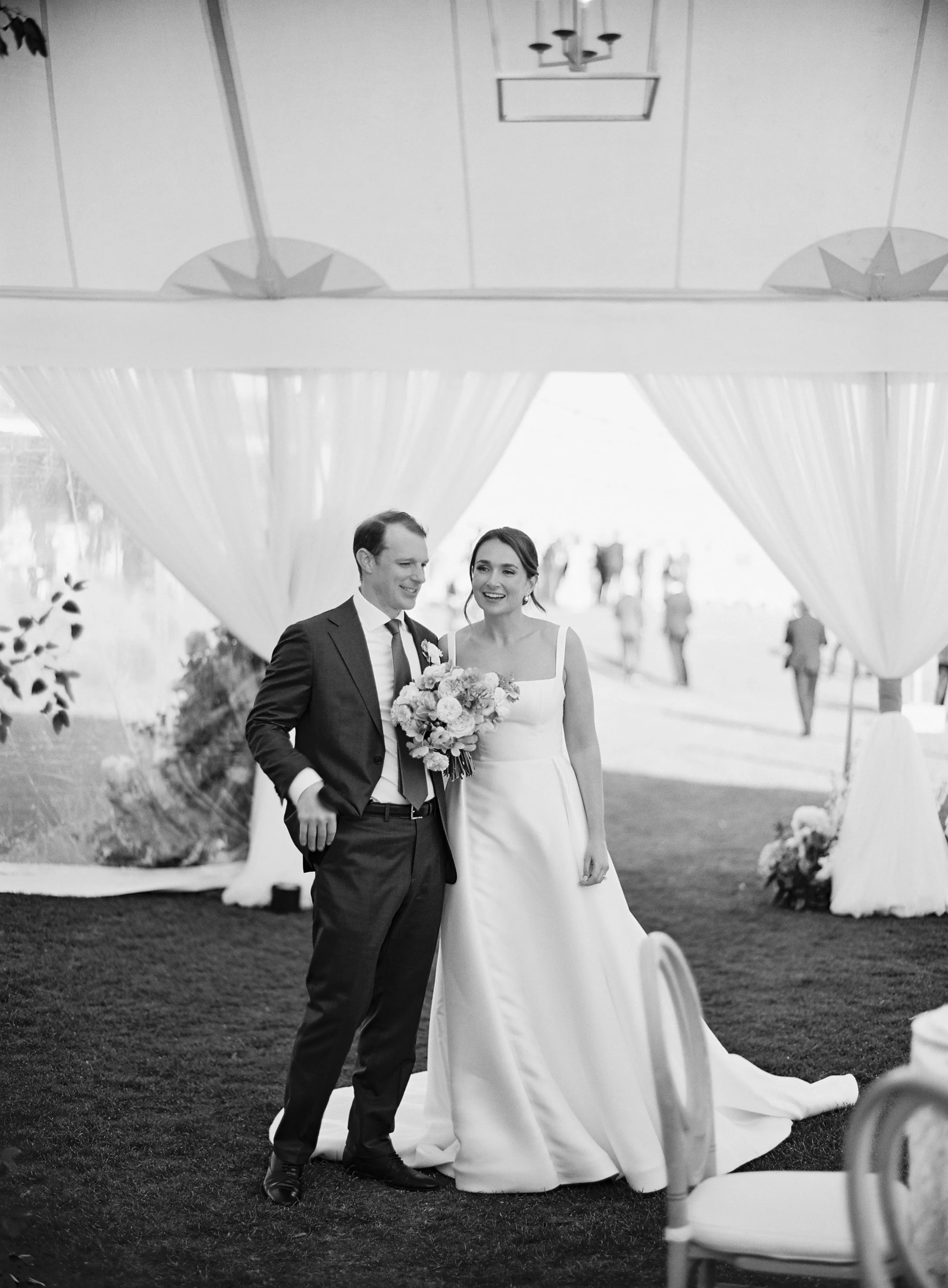 A bride and groom standing together, smiling, with a wedding bouquet. The scene appears to be inside a decorated venue.