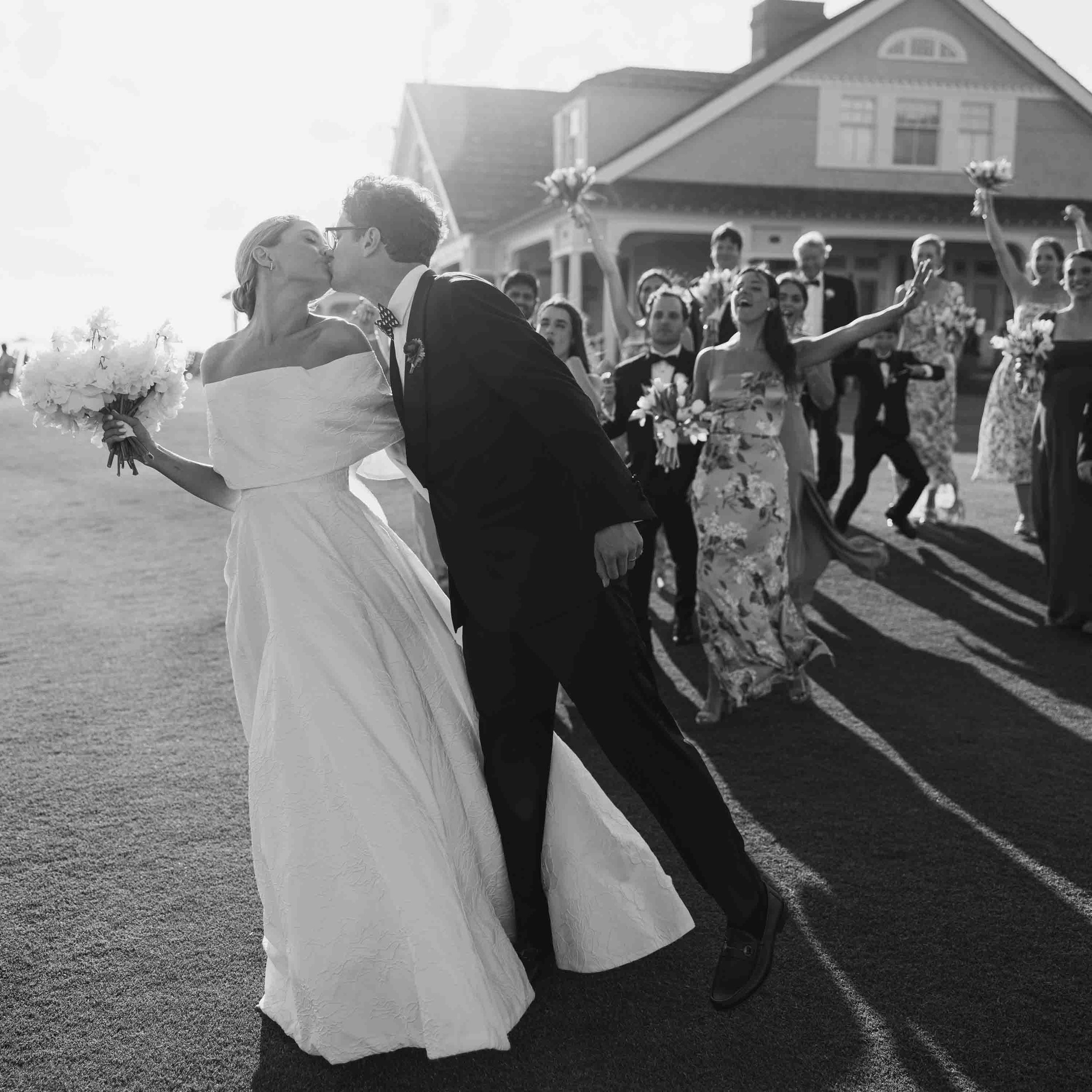 Black and white photo of bride and groom kissing after their ceremony with guests celebrating behind them