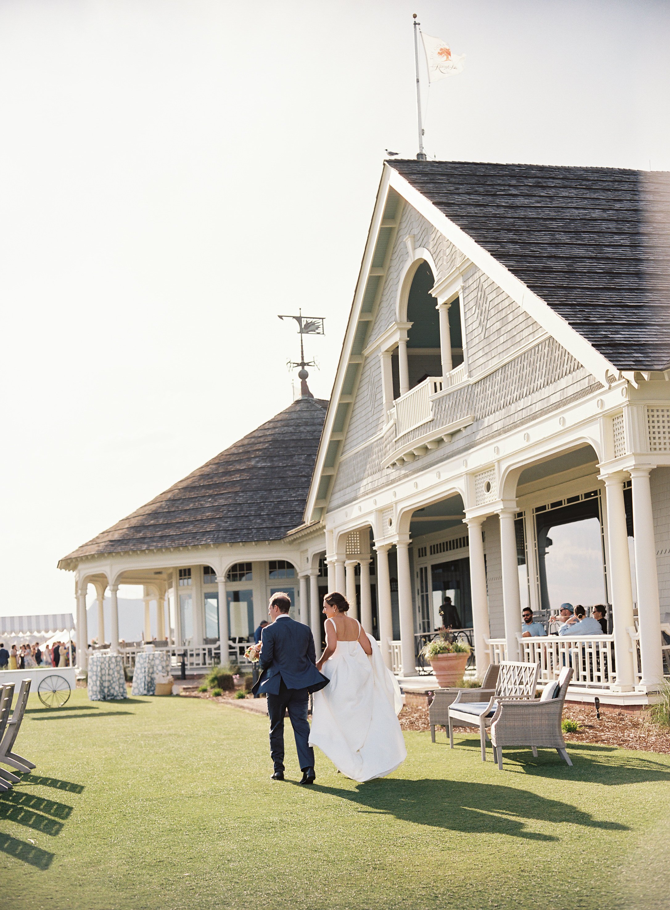 A bride and groom walking away from a large, white, Victorian-style house with a wraparound porch at a wedding venue. The bride is in a white wedding gown, and the groom is in a blue suit. Guests are seated on the porch, and the setting is sunny with