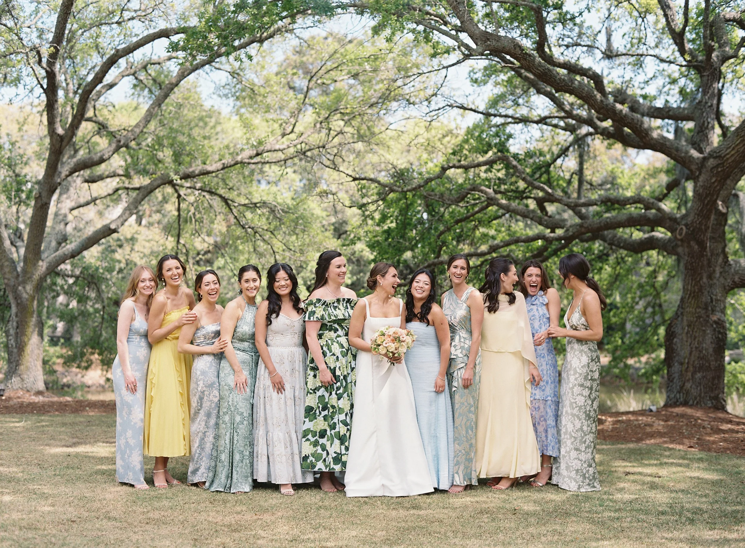 A group of women, including a bride in a white dress holding a bouquet, and her bridesmaids in pastel dresses, standing outdoors on grass under large trees, smiling and laughing.