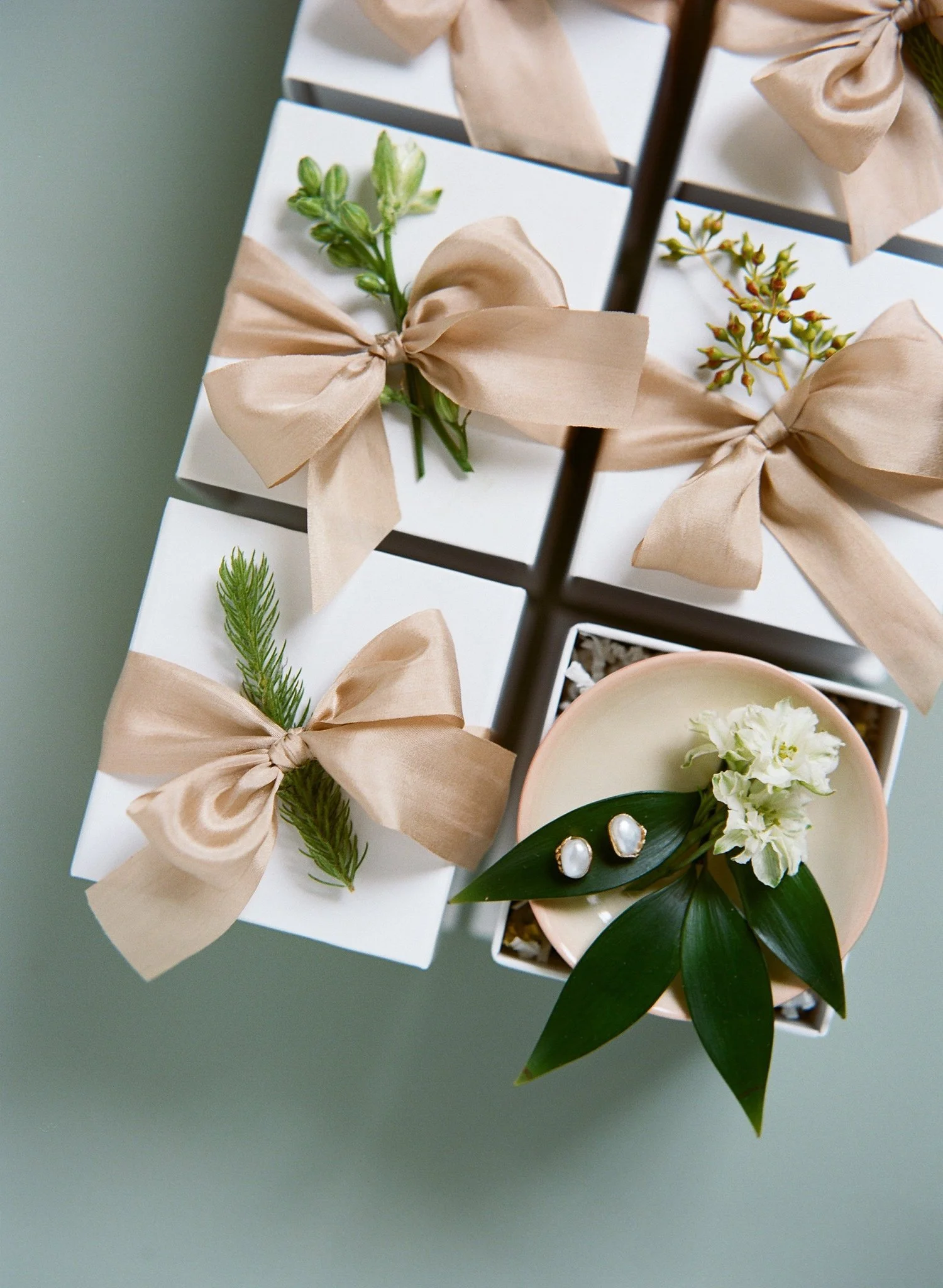 Elegant wedding flat lay featuring white gift boxes tied with beige silk ribbons and greenery, styled with pearl earrings and white florals in a blush bowl.
