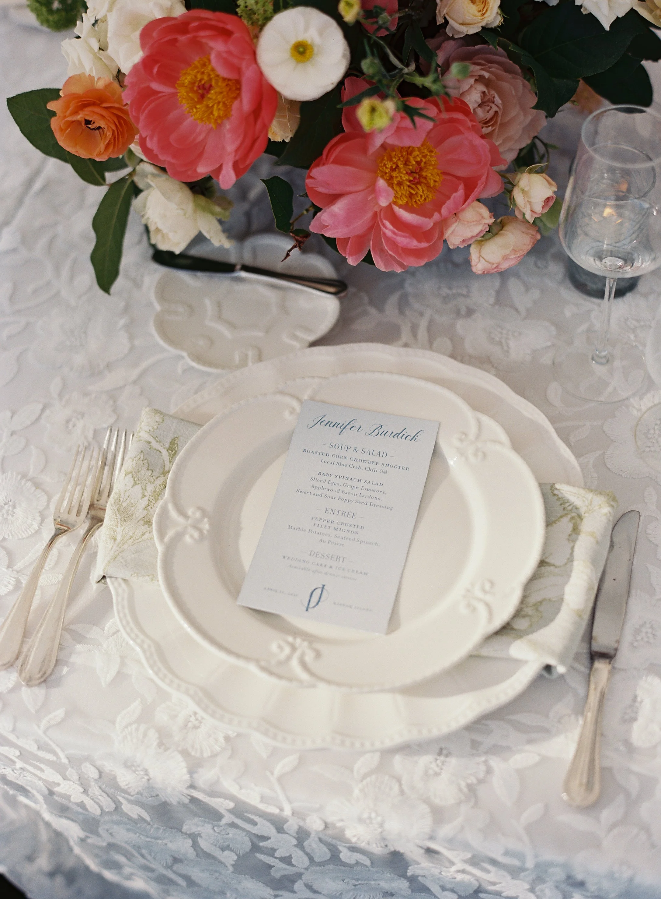 Elegant table setting with a white floral tablecloth, a decorative white plate, a menu card, silverware, a glass, and a large floral centerpiece featuring pink, white, and peach flowers.