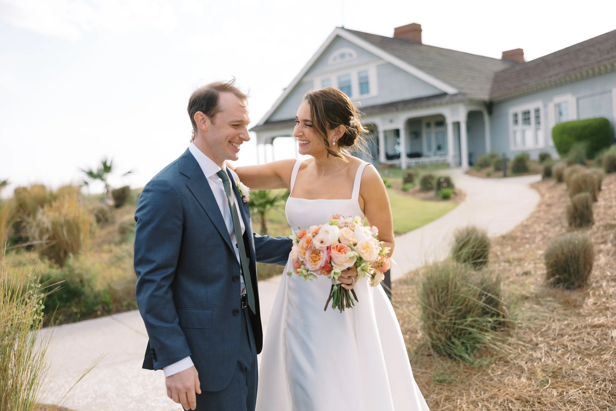 A bride and groom smiling and sharing a moment outside in front of a large house. The bride is holding a bouquet of pink and white flowers, and the groom is wearing a blue suit with a white shirt and tie.