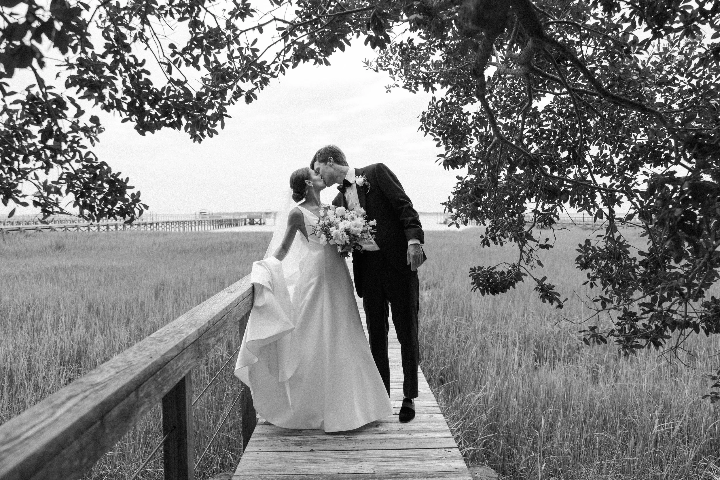 Black and white film photograph of bride and groom kissing on marsh boardwalk in Charleston