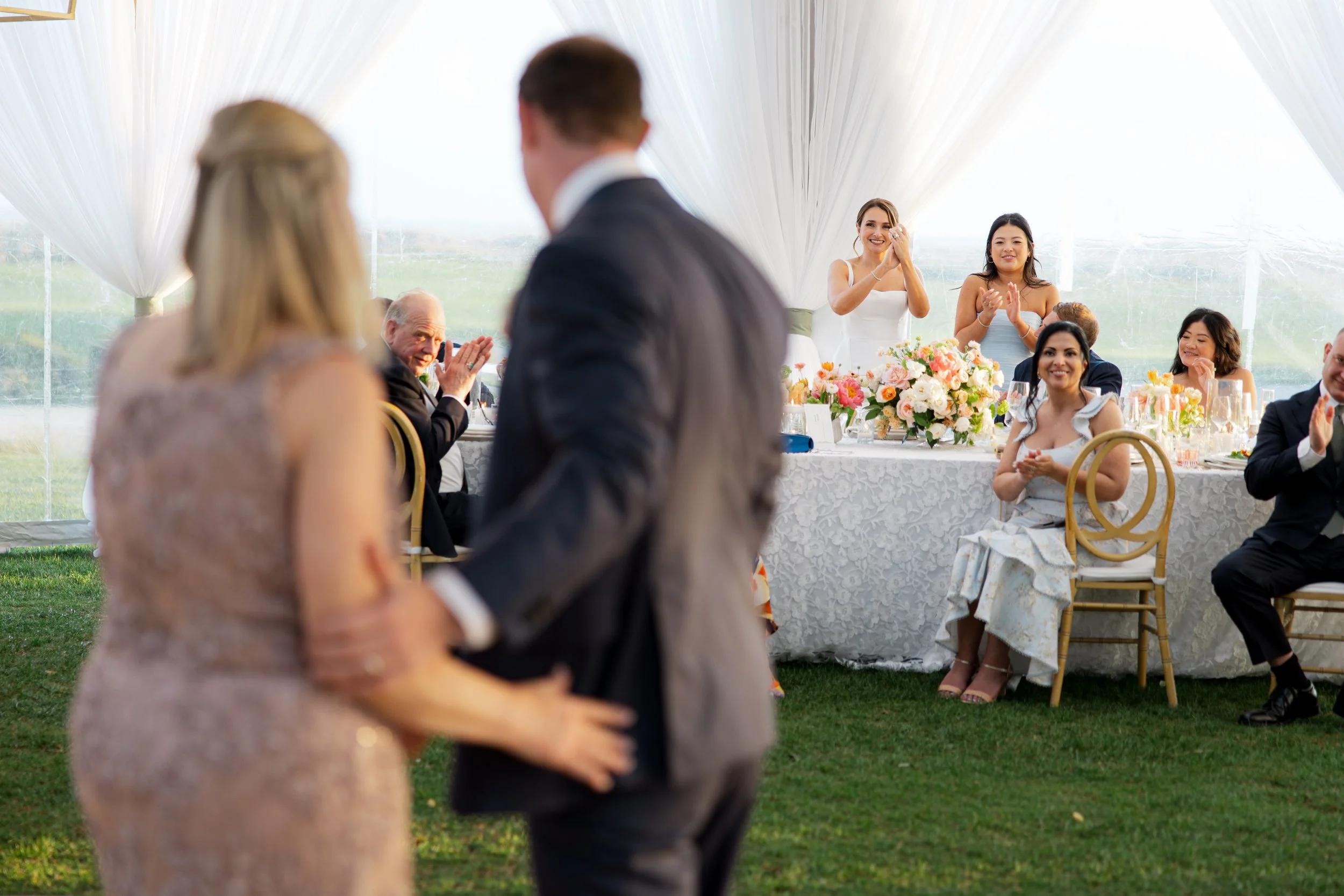 Couple dancing at a wedding reception outside, with guests clapping and smiling at a decorated head table in the background.