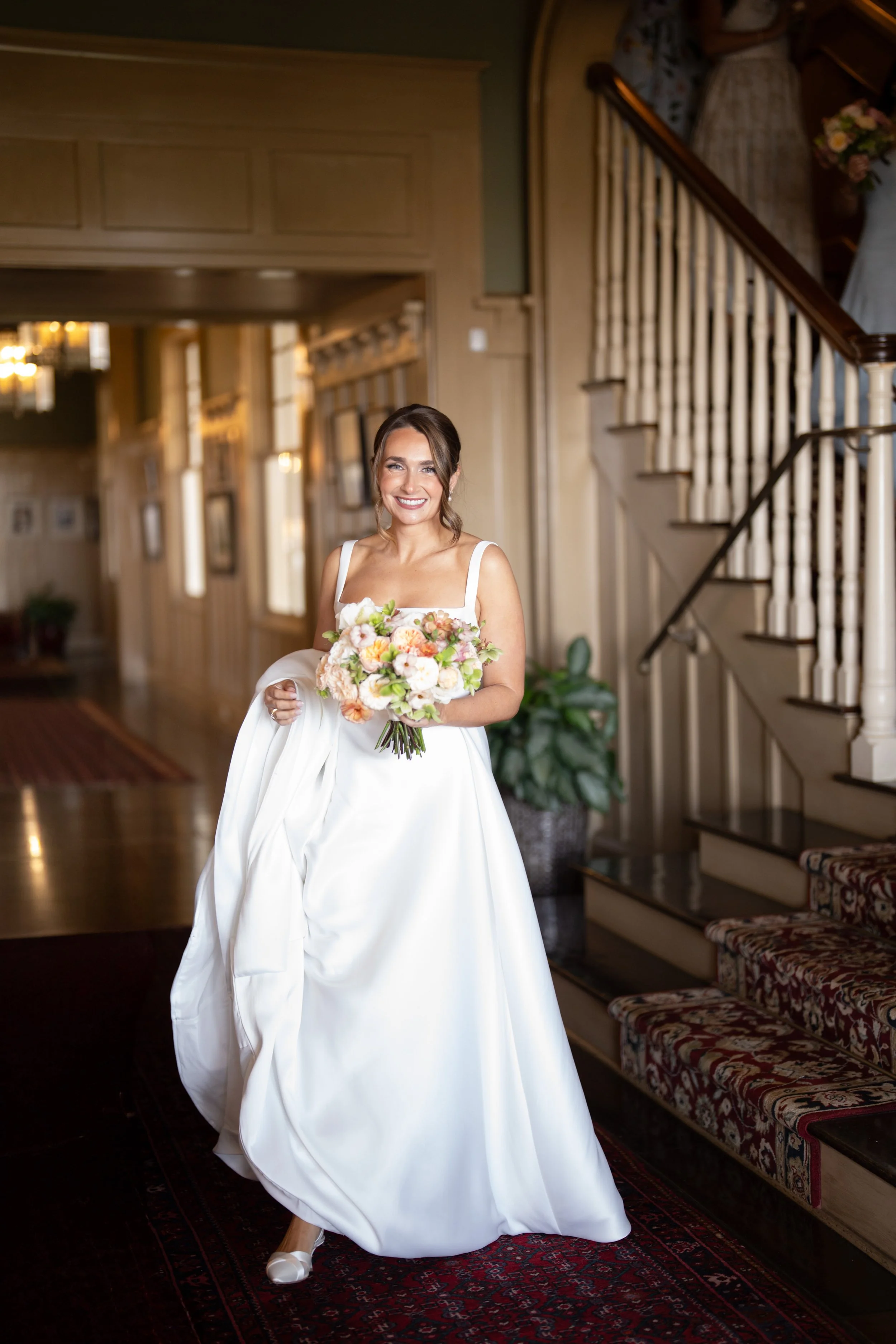 A bride in a white wedding gown holding a bouquet of flowers, walking inside a historic or elegant venue with wooden stairs and decorative decor.