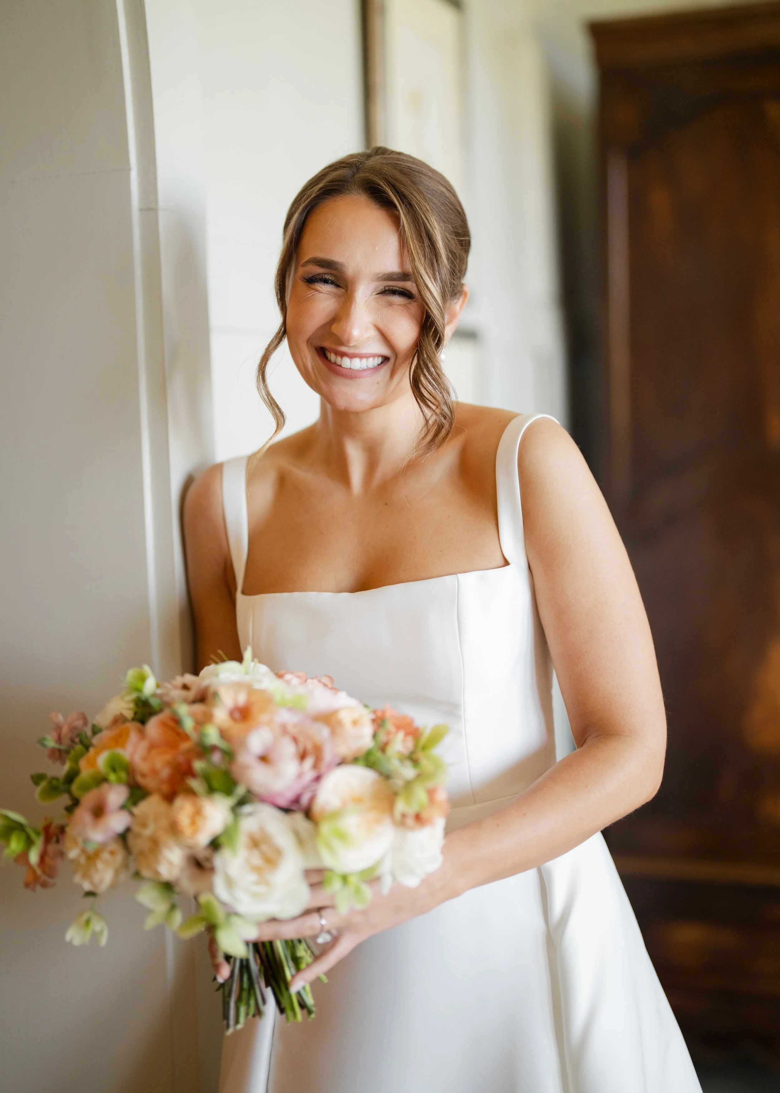 A woman in a wedding dress holding a bouquet of flowers, smiling, standing indoors near a wall.