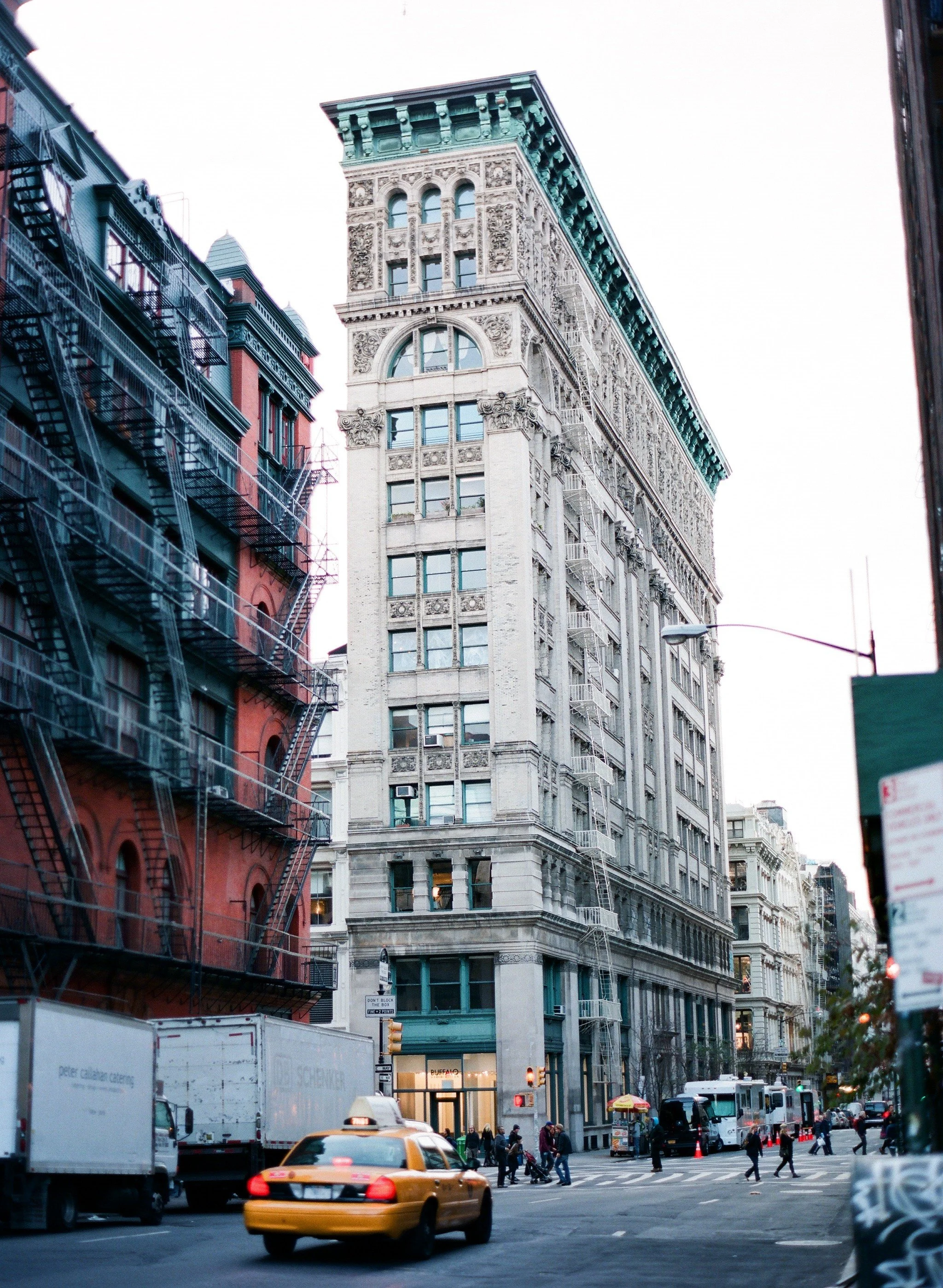 A city street scene with a tall, ornate beige building in New York, a yellow taxi in motion, pedestrians crossing the street, and a red brick building with fire escapes on the left.