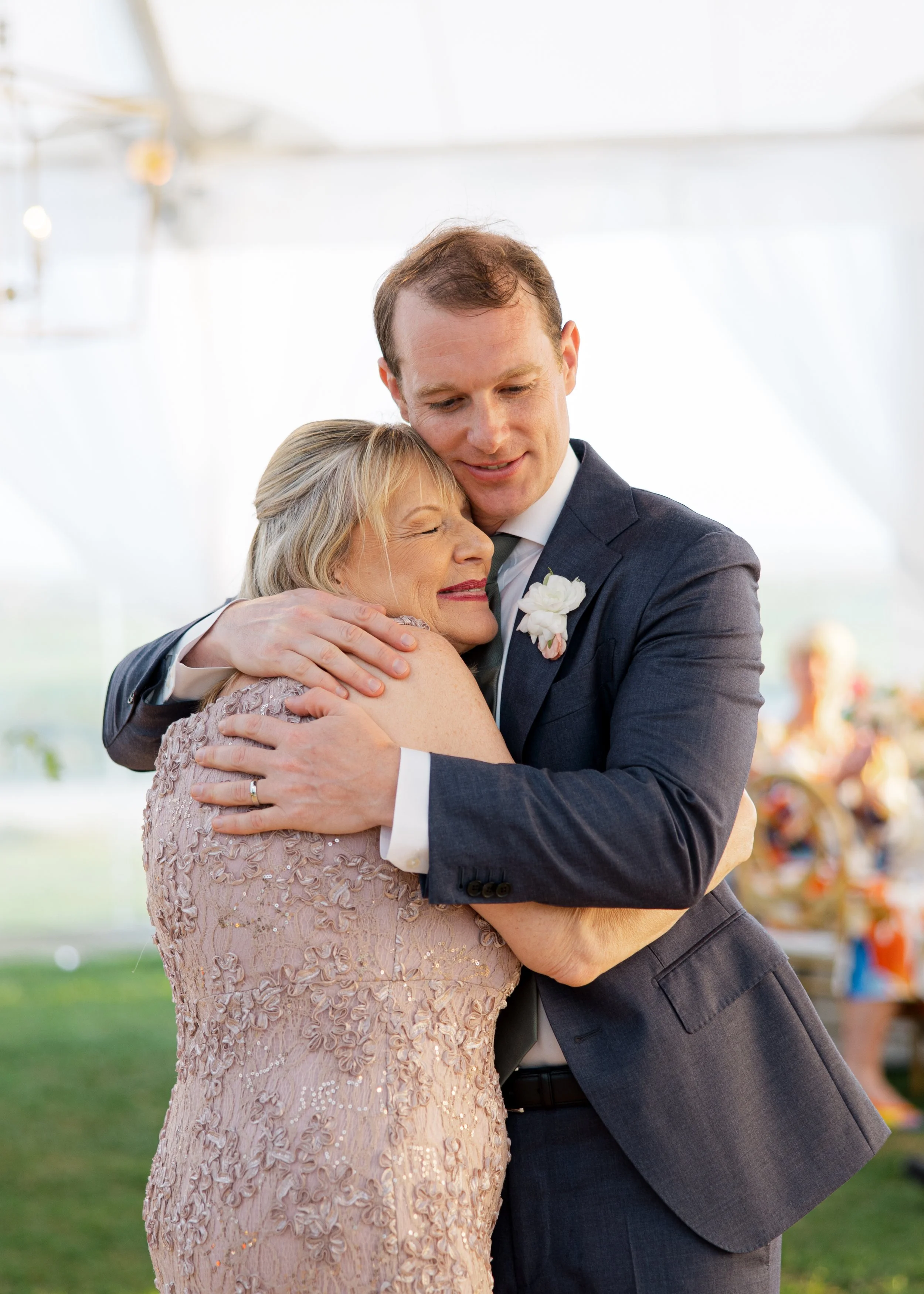 A young man in a suit hugging an older woman in a pink lace dress at a wedding reception.