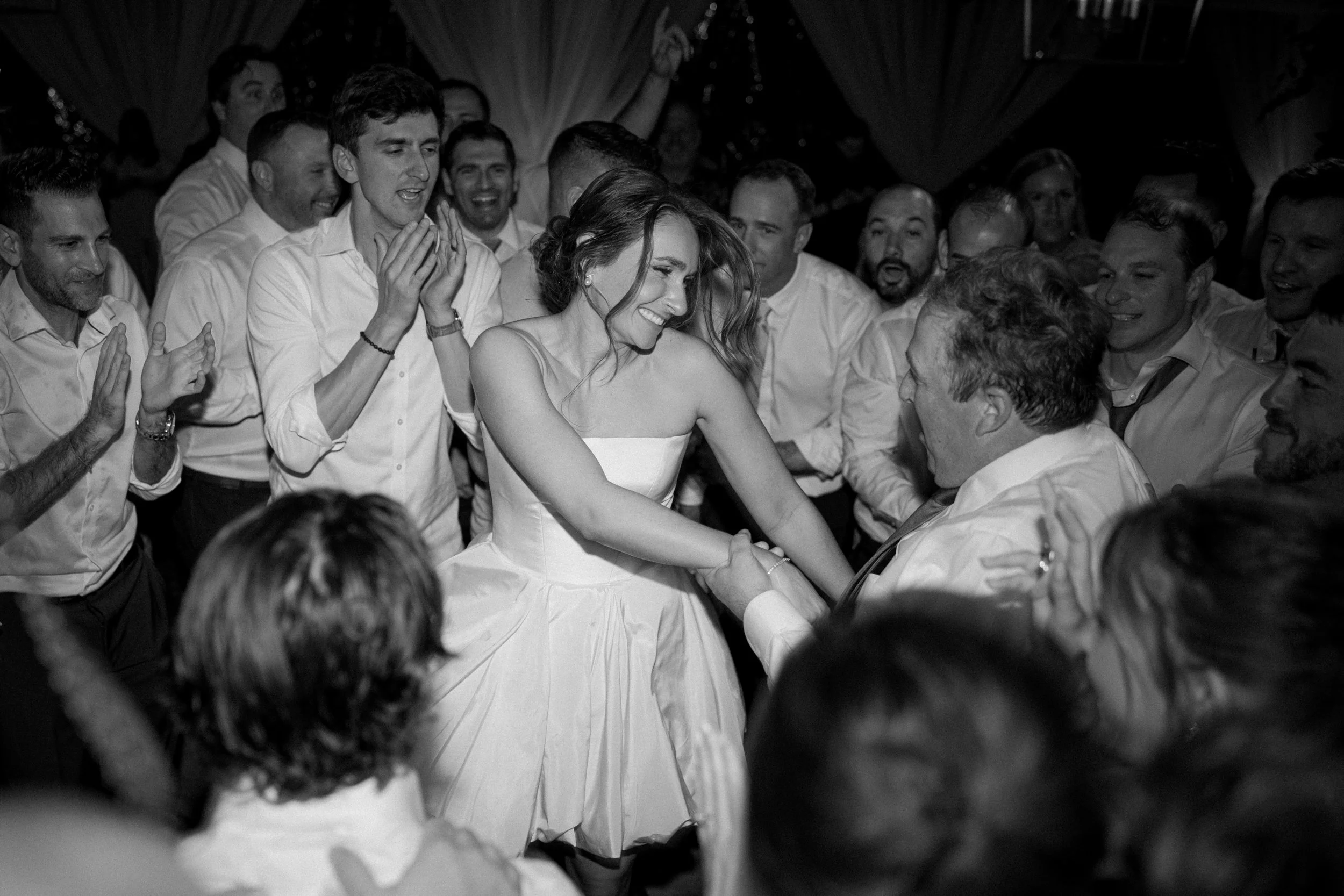 A woman in a sleeveless dress dancing and smiling with a man while surrounded by many men in white shirts at a celebration.