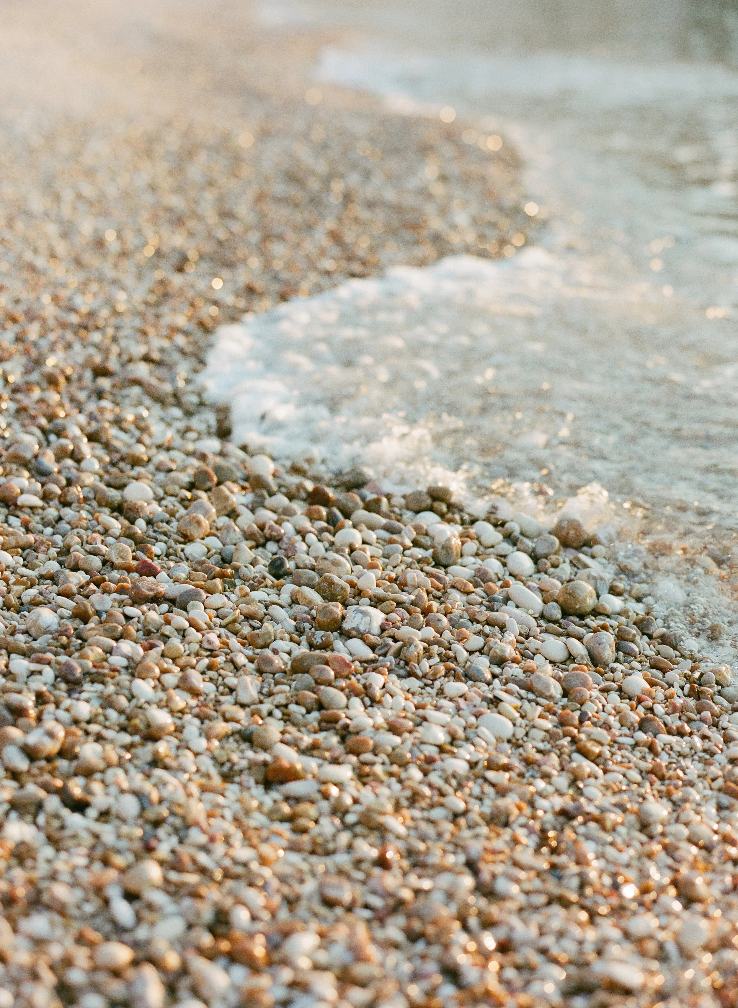 Close-up of a pebble beach in Bari, Italy, with small, smooth stones and gentle waves lapping at the shore, illuminated by soft golden sunlight.