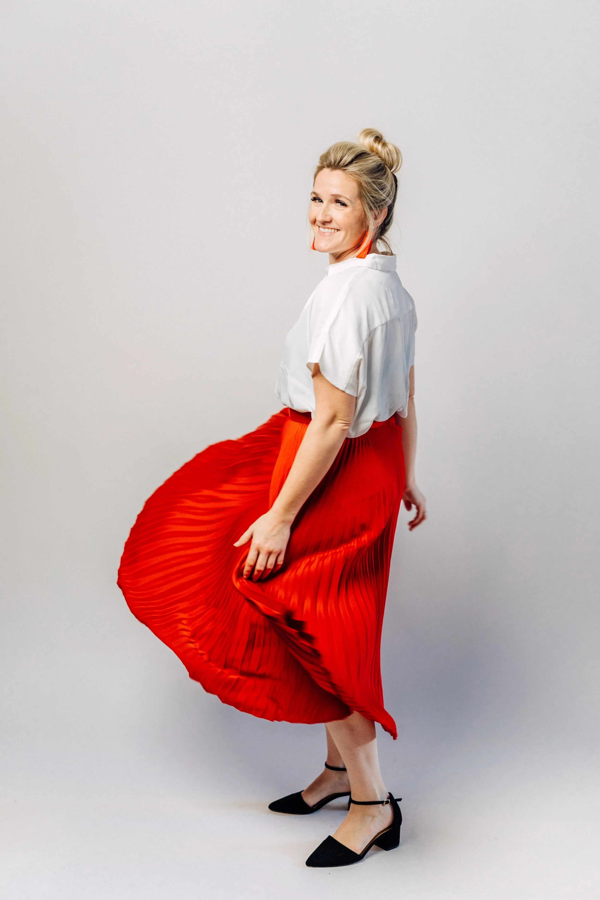 Portrait of Lauren Jonas wearing a white blouse, a red pleated skirt, and black heels, smiling and flicking her red skirt.