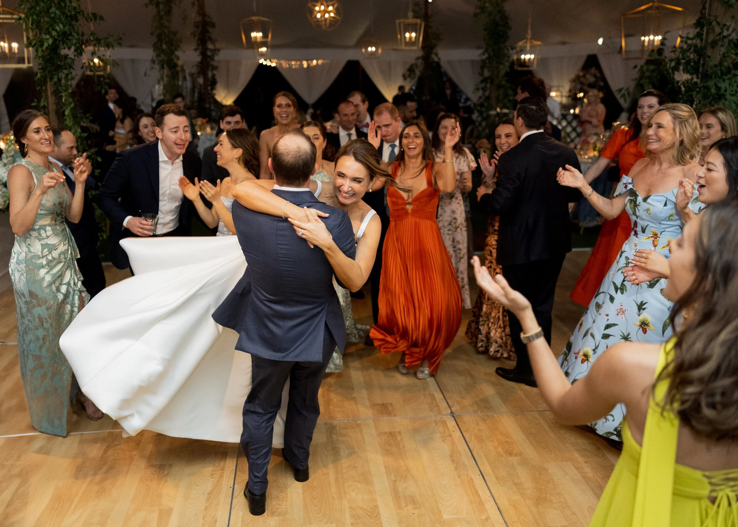 People dancing and celebrating at a wedding reception inside a decorated tent, with a bride in a white wedding dress and guests in colorful dresses and suits.