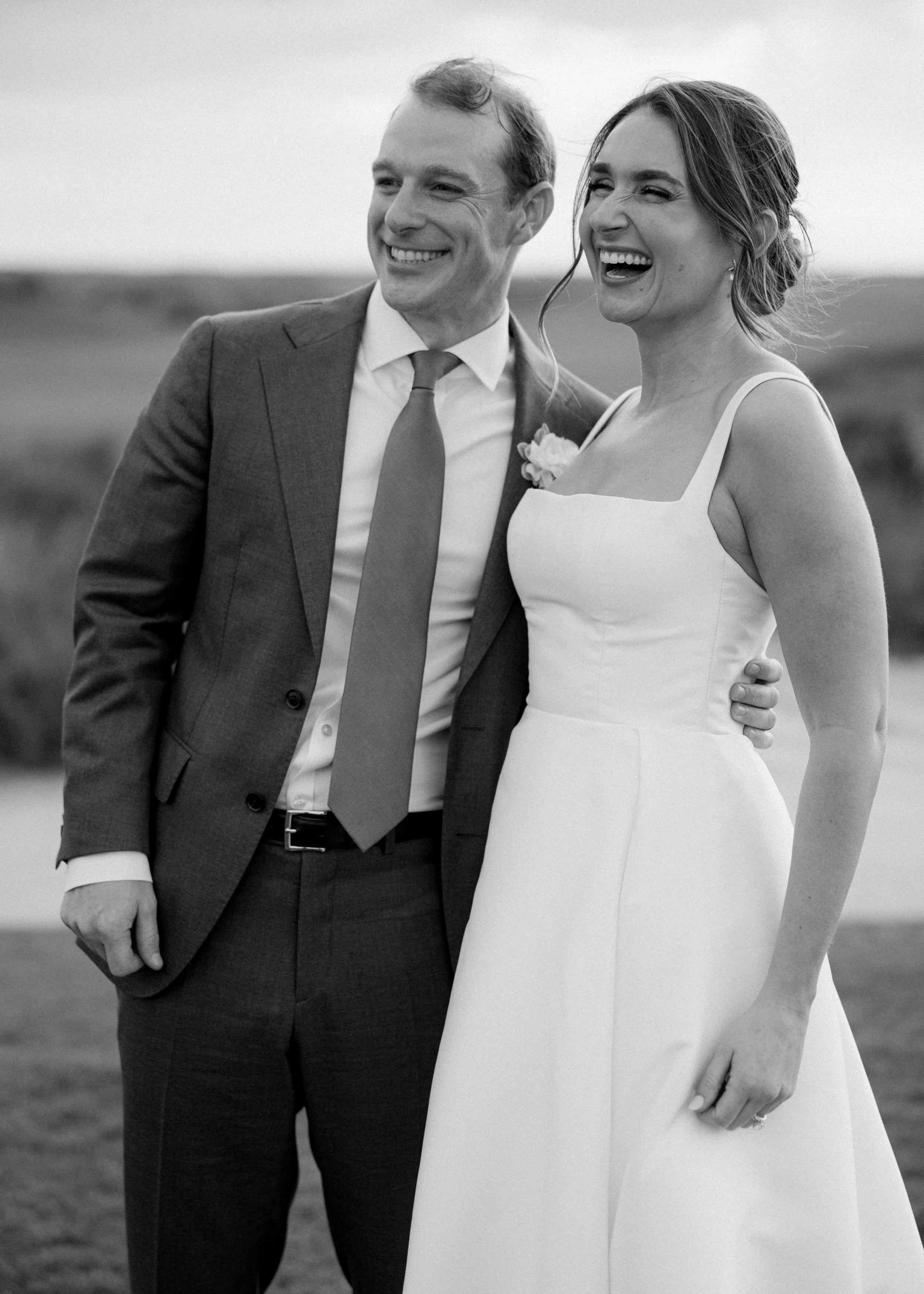 Black and white photo of a smiling couple in wedding attire outdoors, standing close together with arms around each other.