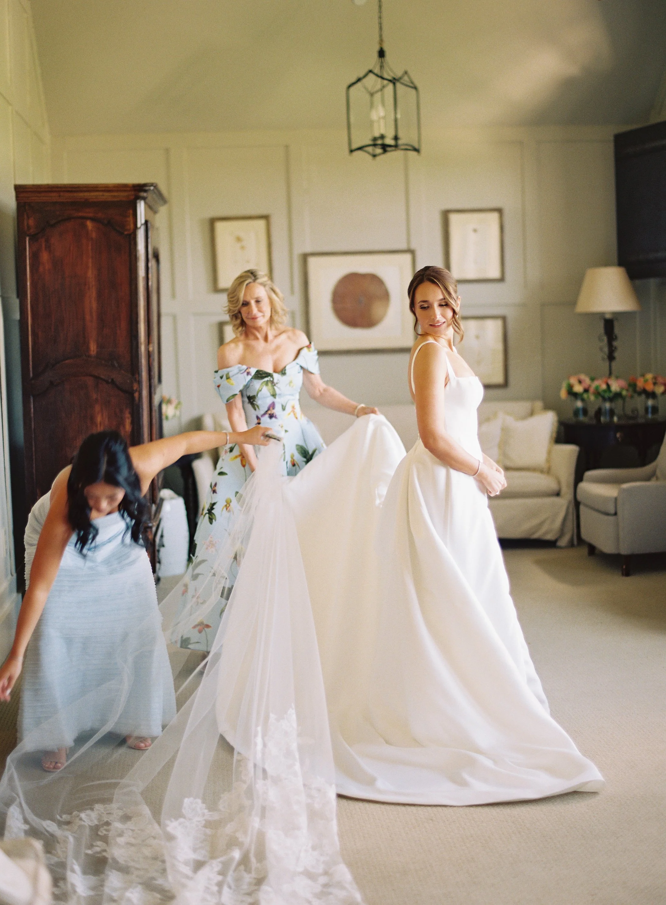 A bride in a white wedding gown stands in a room, as a woman adjusts her dress and a girl in a striped dress with a veil moves nearby.