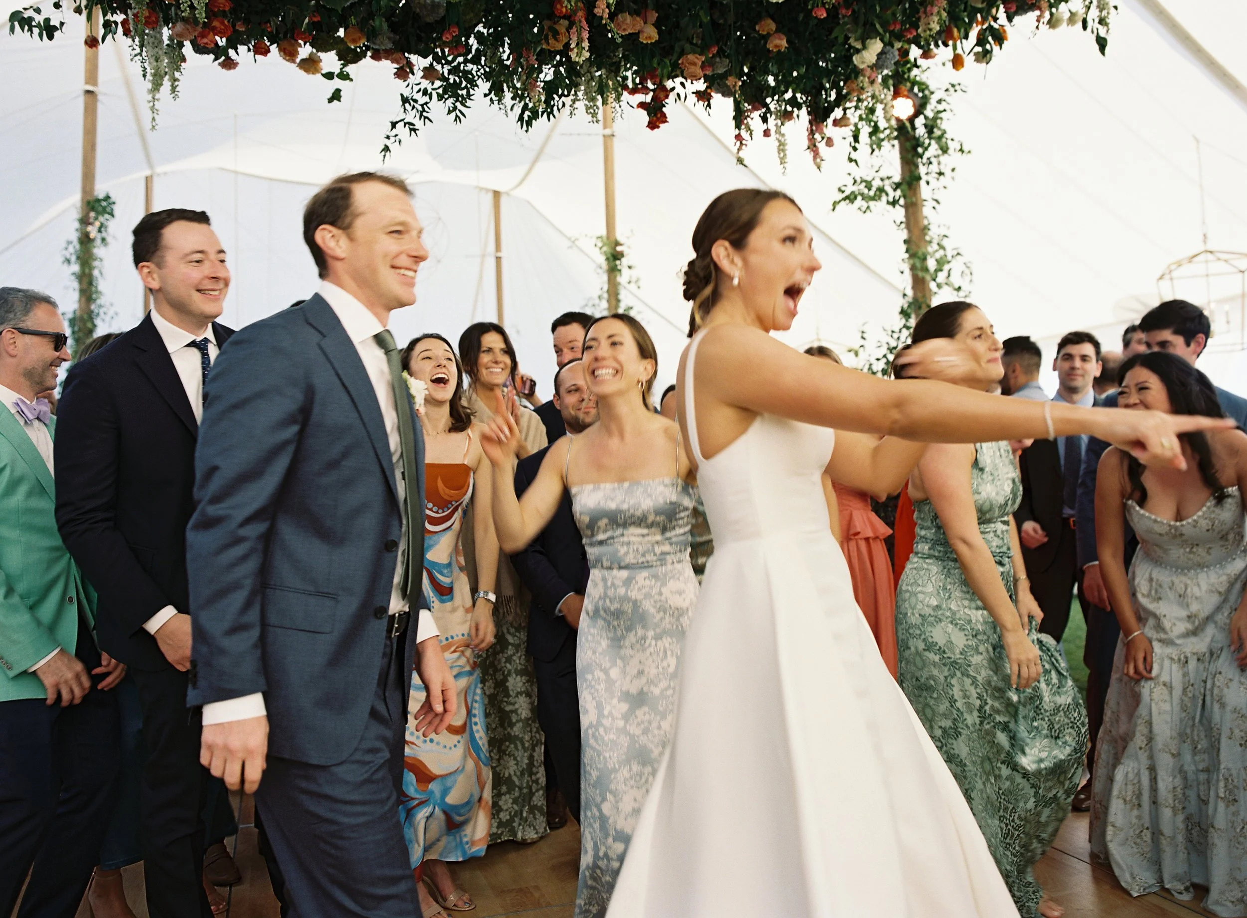 People dancing and celebrating at a wedding reception in a decorated tent, with the bride in a white dress and the groom in a suit.
